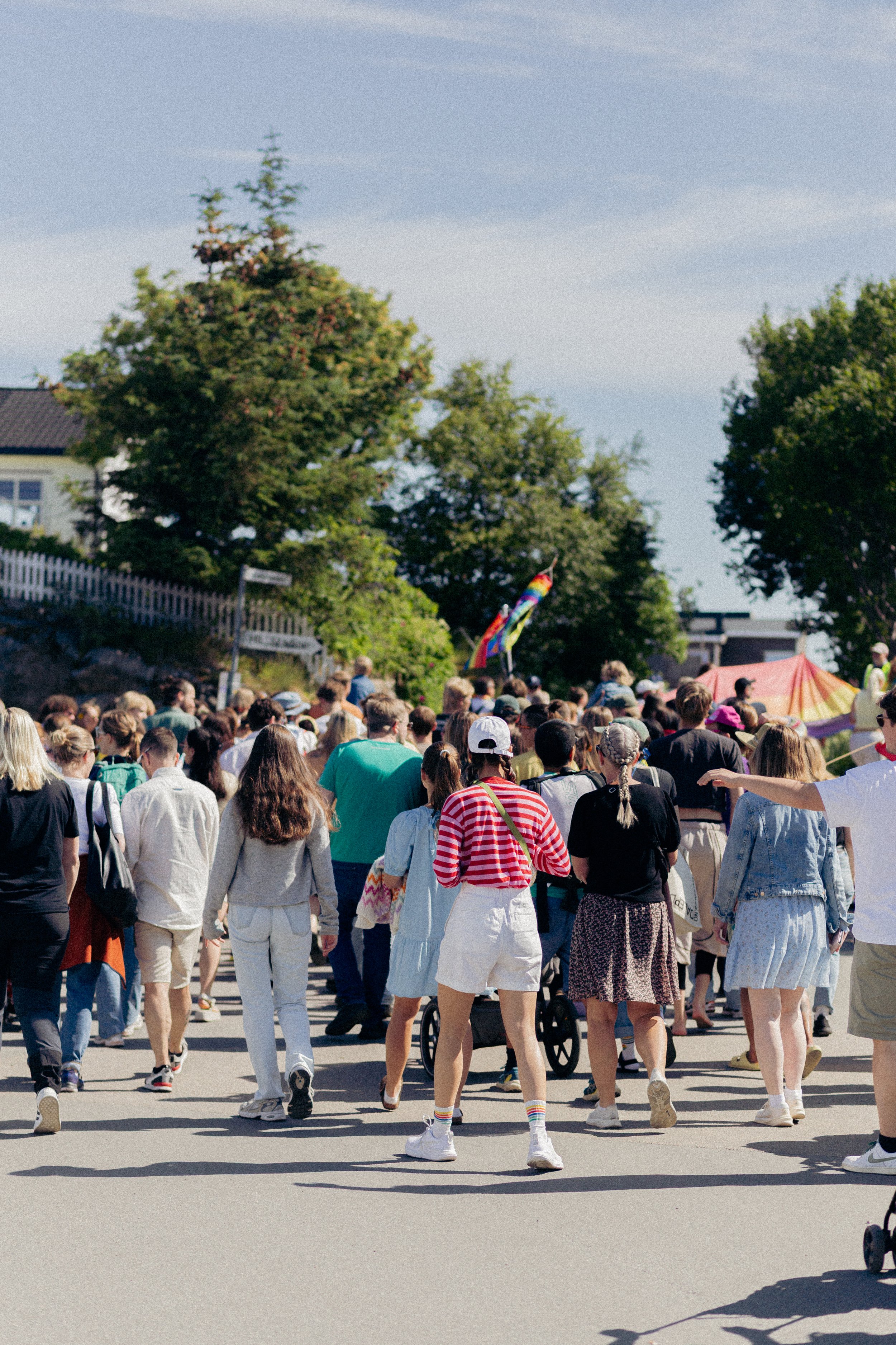 Mennesker samlet i en stor gruppe utendørs, noen holder en rainbow pride-flagg, i en solrik dag med trær og bygninger i bakgrunnen.