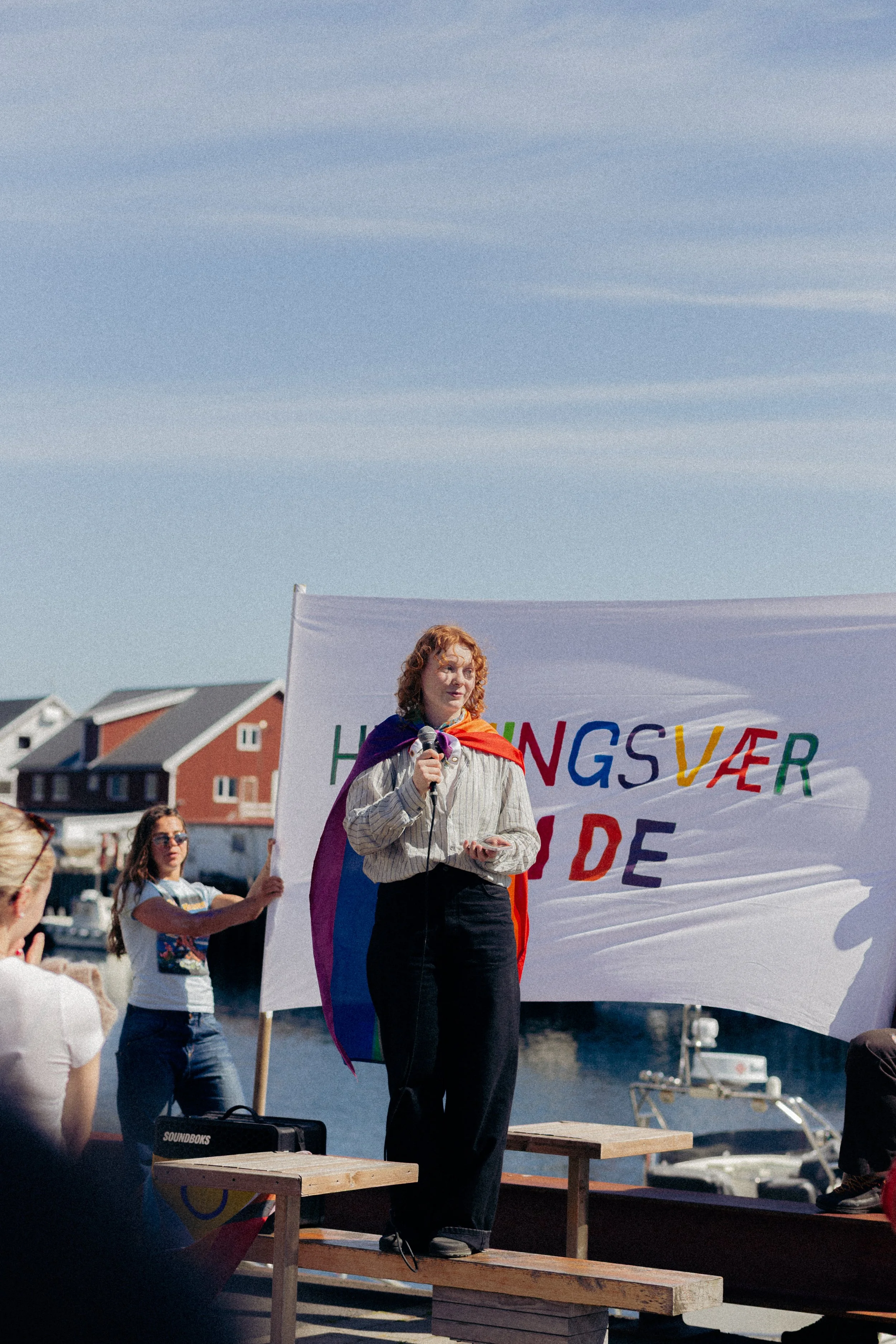 En kvinne holder en mikrofon og taler under en utendørs demonstrasjon. Hun har rød, krøllete hår og er kledd i en stripete skjorte og svarte bukser. Bak henne holder flere personer opp et stort farget banner med tekst, og situasjonen er ved et vann med båter og hus i bakgrunnen.