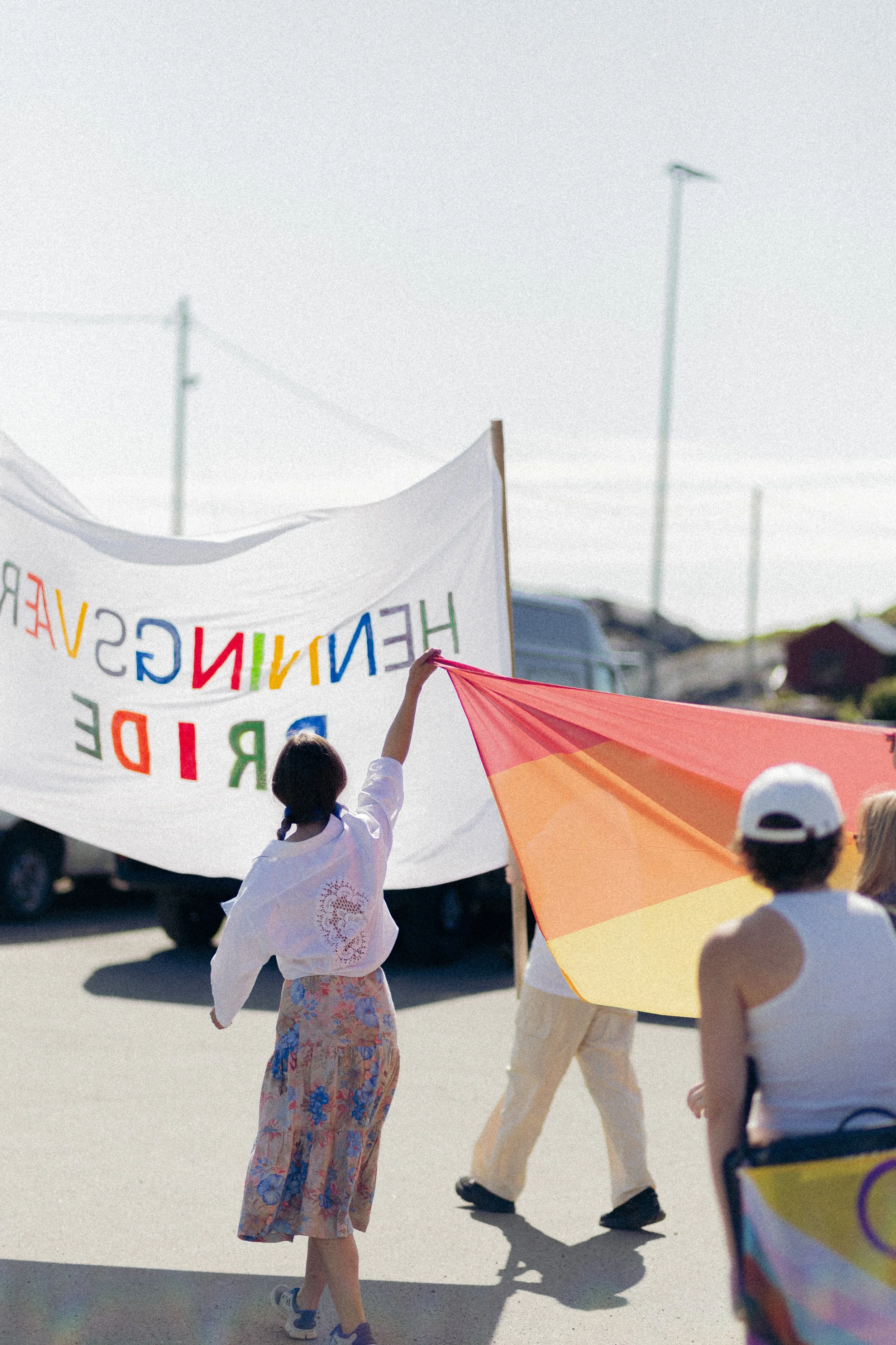 Barn og unge som holder et stort fargert banner og en regnbuetelt under en parade eller marsj på åpen gate, med flere personer i bakgrunnen og gatelamper.