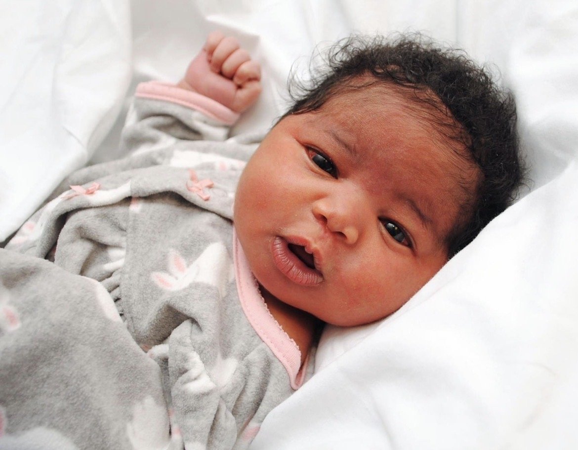 A newborn baby lying on a white surface, wearing a gray outfit with pink accents, with dark curly hair and open eyes.