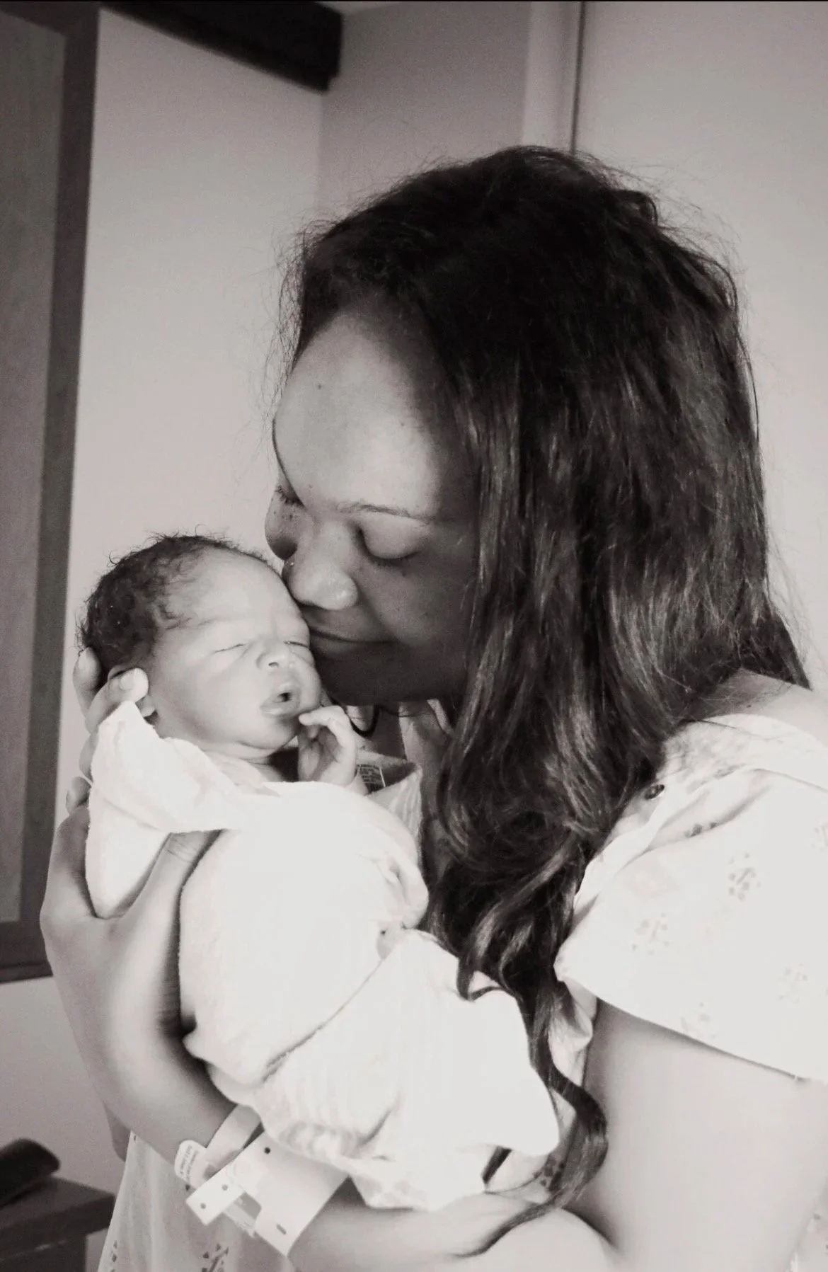 A woman holding a newborn baby close, kissing the baby on the forehead in a hospital setting.