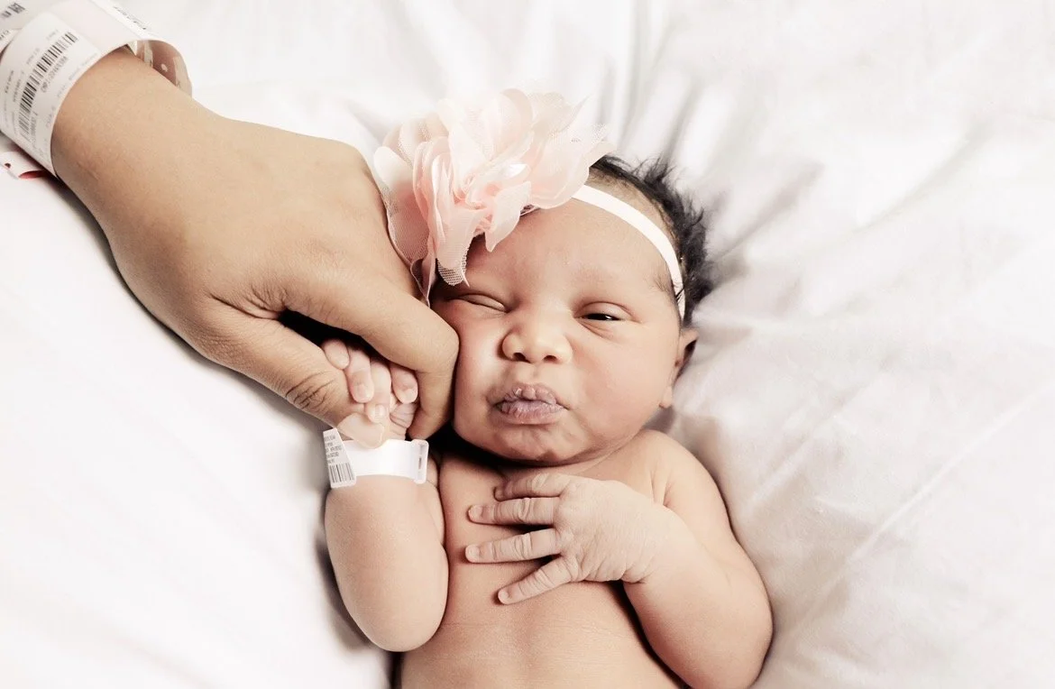 A newborn baby lying in a hospital bed, holding an adult's finger with one hand, with a pink floral headband on her head, making a scrunched-up face.