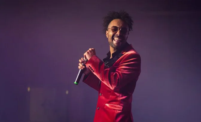 Man with curly hair and sunglasses holding a microphone on stage, smiling, wearing a red blazer and black shirt.