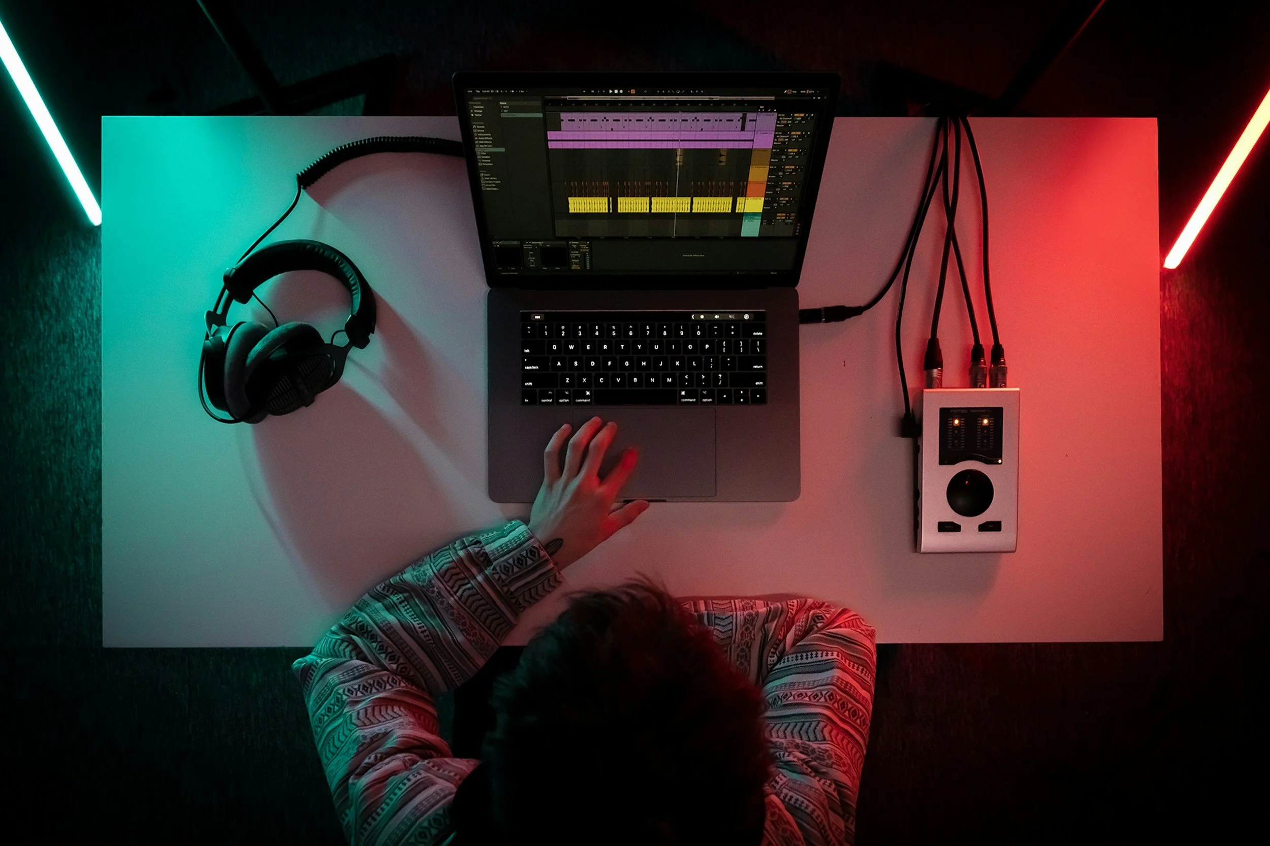 Top-down view of a person working on music production on a laptop with a digital audio workstation open. The workspace includes a pair of headphones, a speaker, and LED strip lights emitting blue and red lighting.
