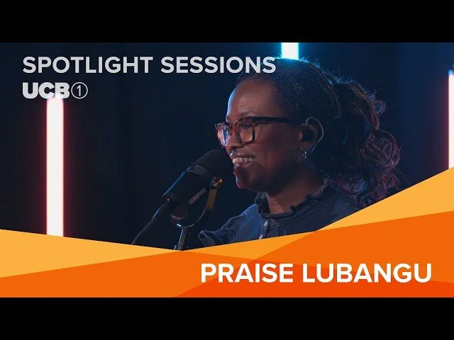 A woman with glasses and long curly hair speaking into a microphone during a panel or interview setting, with a dark background and colorful lighting, labeled 'Spotlight Sessions UCB, Praise Lubangu'.