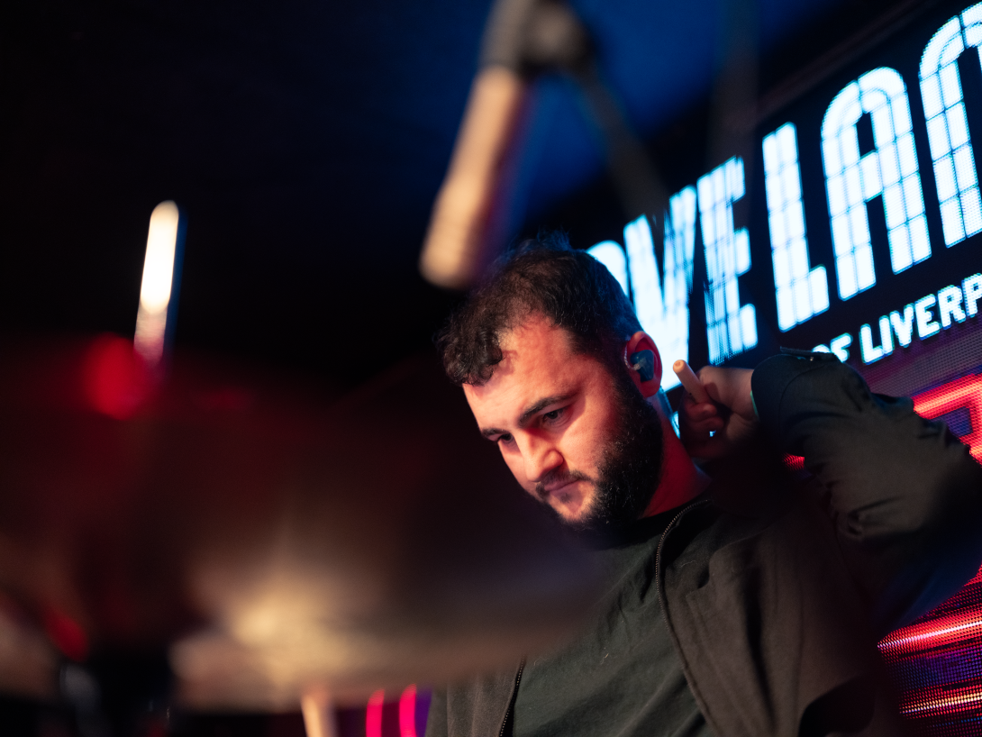 A man with dark hair and a beard adjusts his in-ear monitor while standing in a venue with bright, colorful LED lights behind him.