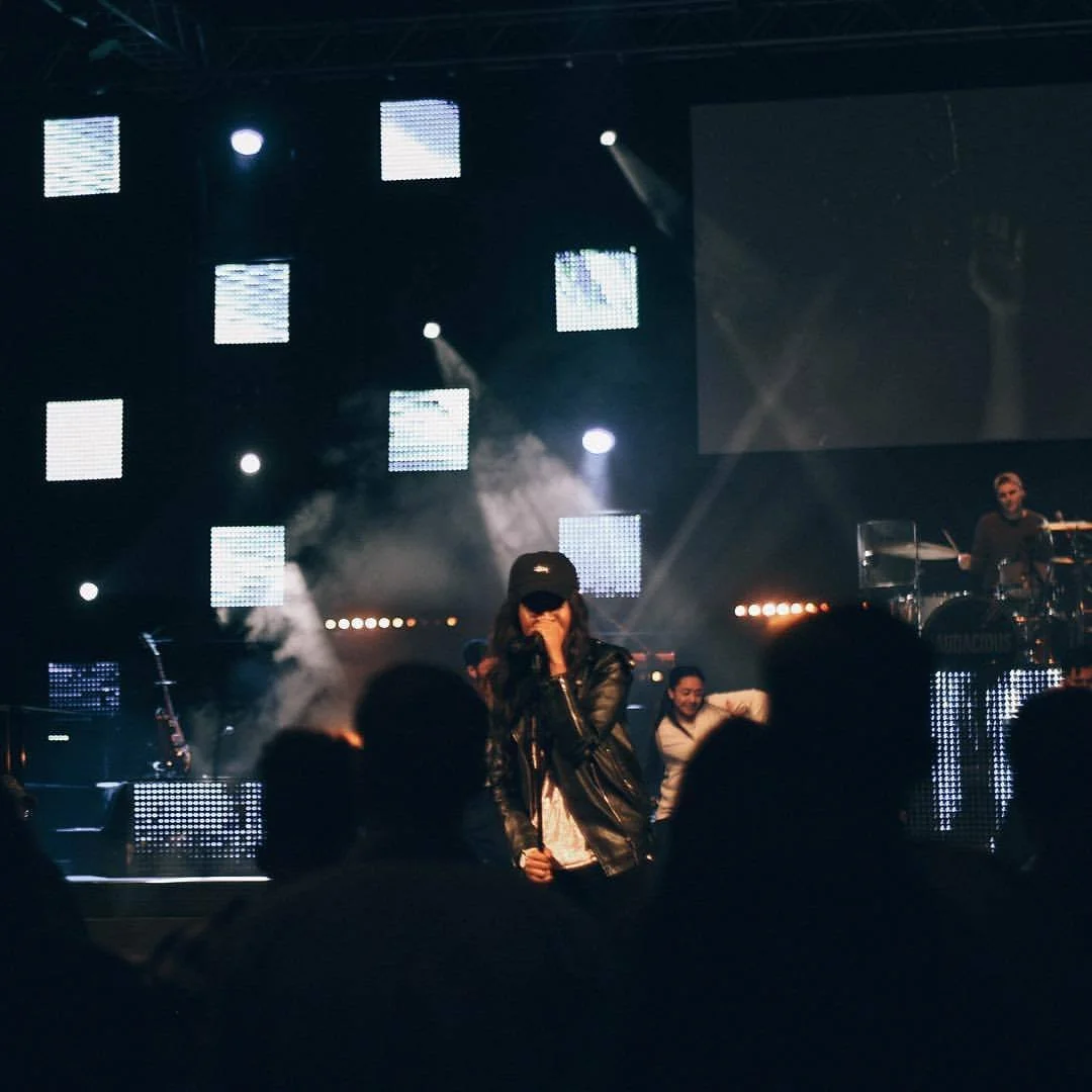 A musical concert with a female singer on stage wearing a black leather jacket and baseball cap, surrounded by band members and illuminated by bright stage lights.