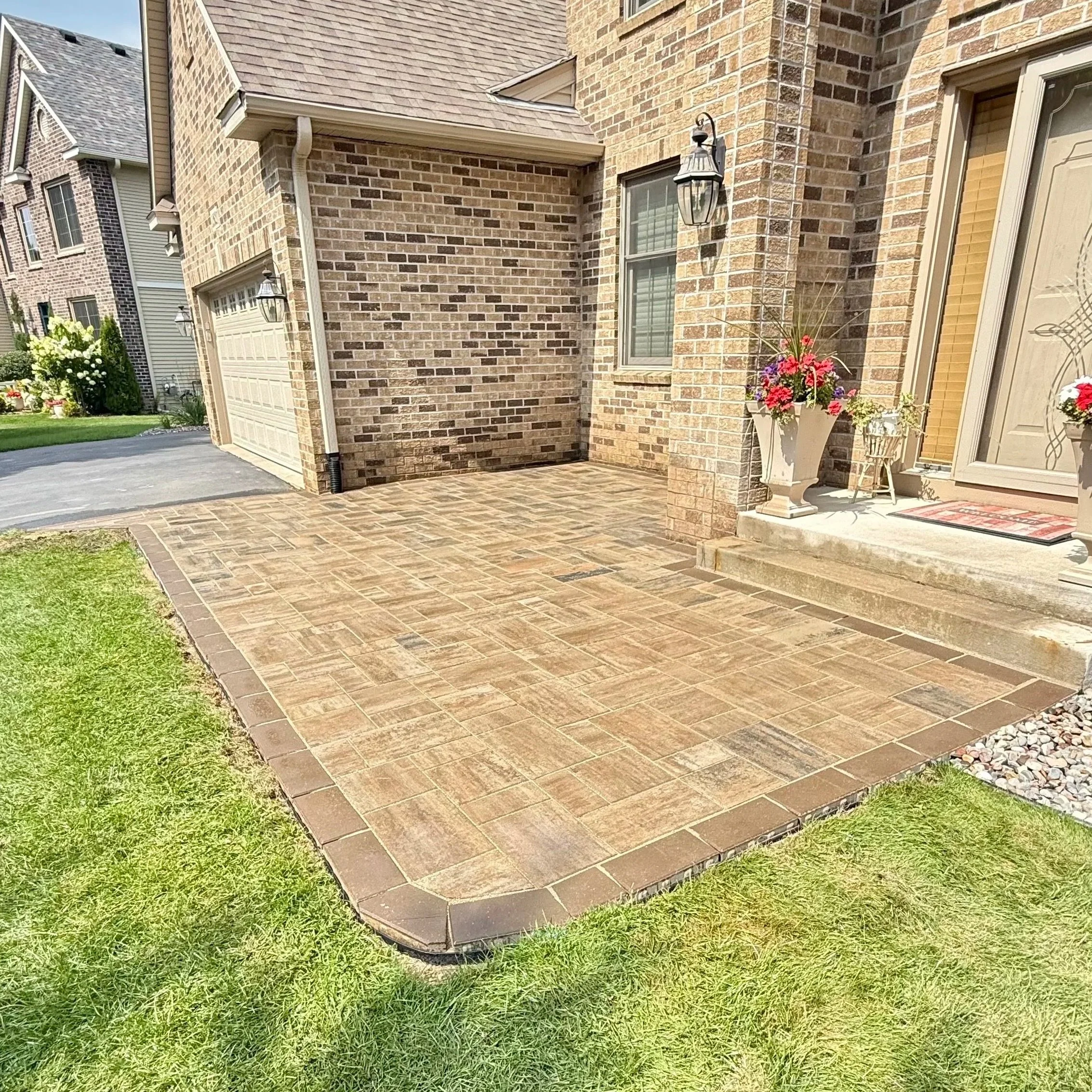 A brick house exterior with a garage, a paved driveway, a flowerpot with pink and purple flowers, and a patio.