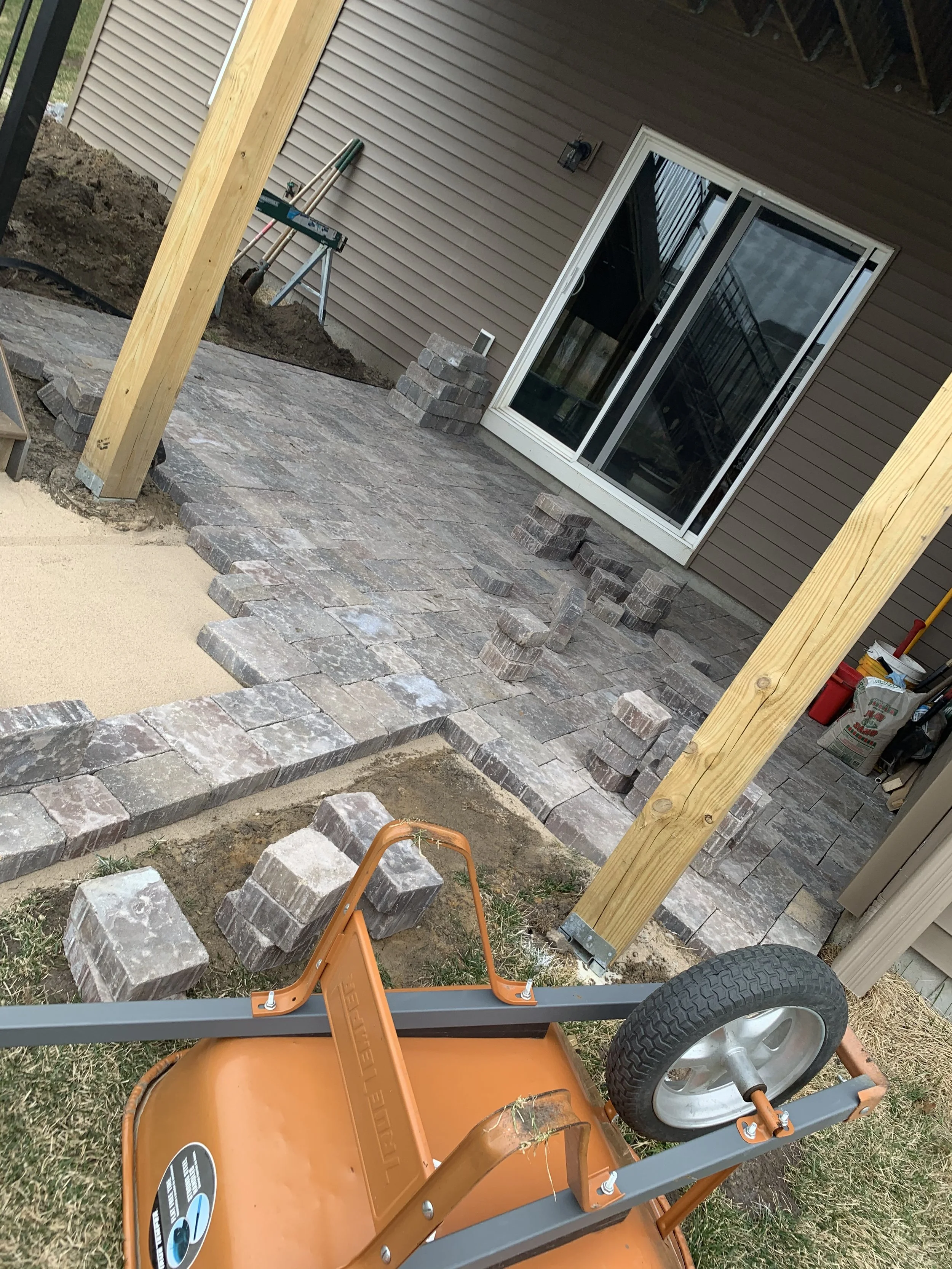 A construction site with a partially completed brick patio, wooden posts, and construction tools including a wheelbarrow, shovels, and a saw. The patio is adjacent to an outdoor house area with a sliding glass door and beige siding.