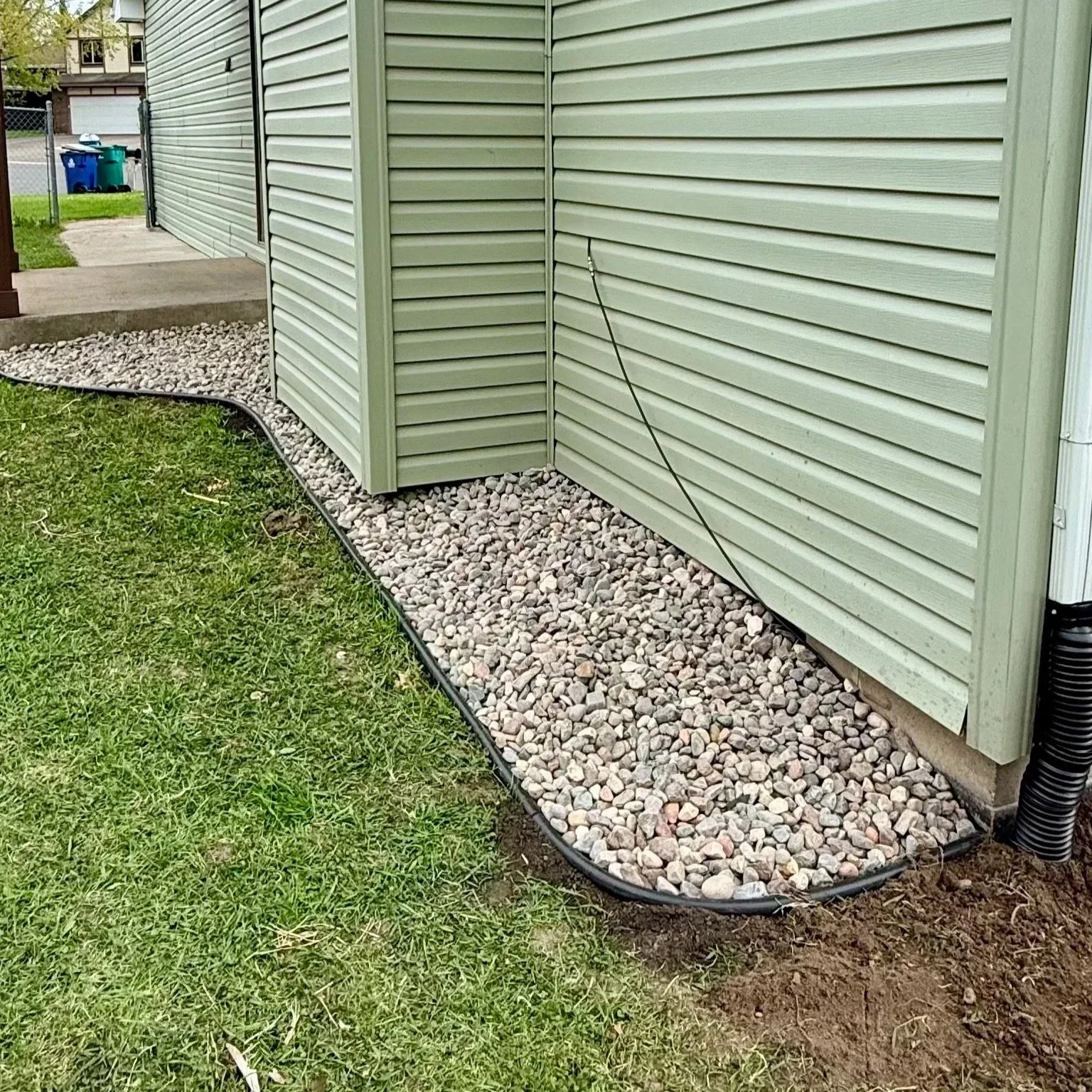 The corner of a house with light green vinyl siding, a gravel bed edged with black trim, green grass, and a black drain pipe on the right side.