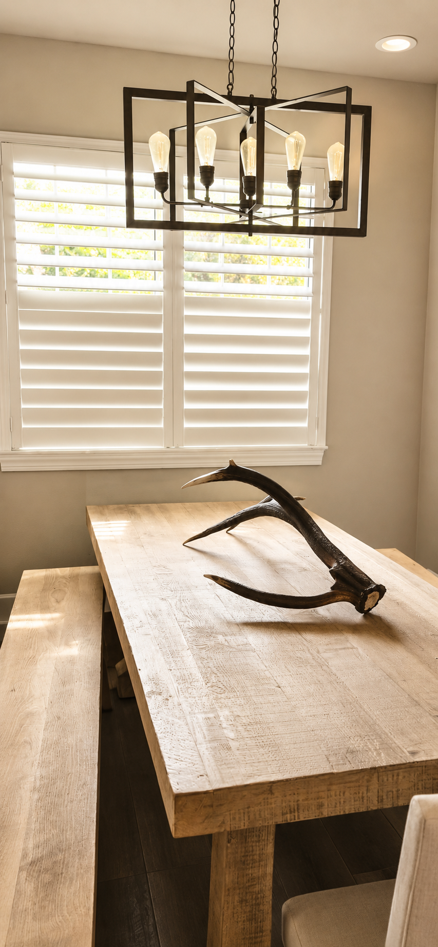 Rustic wooden dining table with antlers on top, near a window with white plantation shutters, and a modern black chandelier with exposed bulbs hanging above.