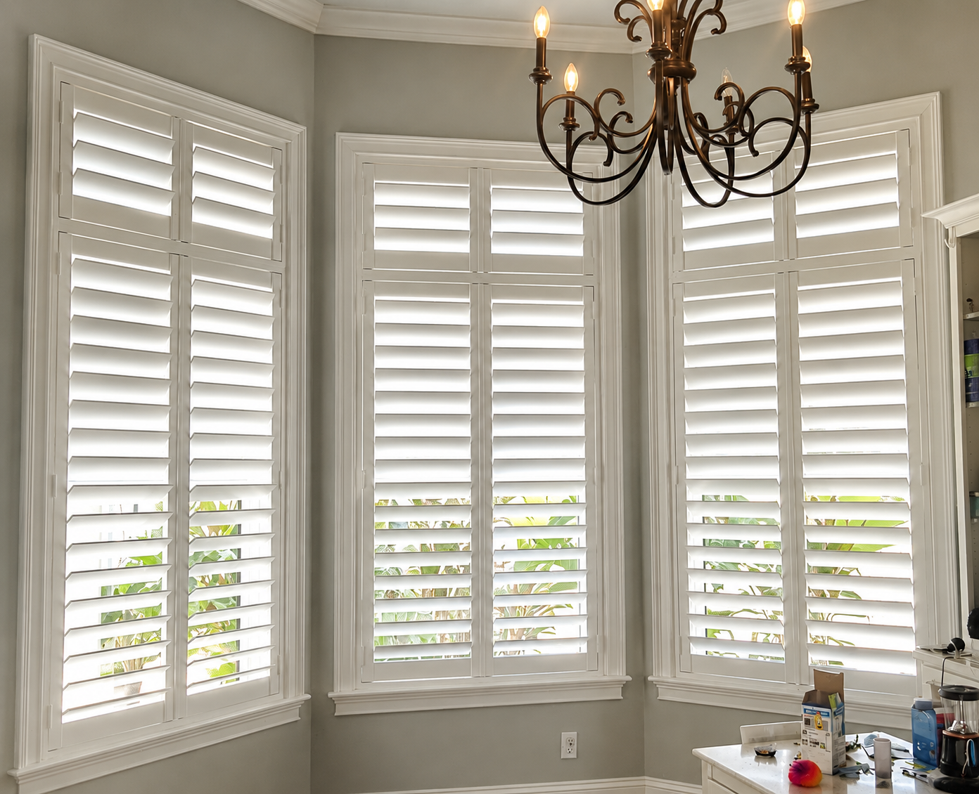 A corner of a room with large white plantation shutters on three windows, a chandelier hanging from the ceiling.