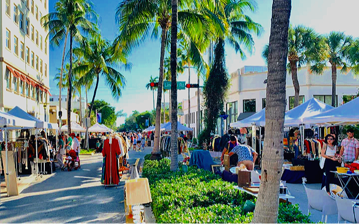 Outdoor street market with white tents, vendors, shoppers, palm trees, and bright sunny weather in a downtown area.