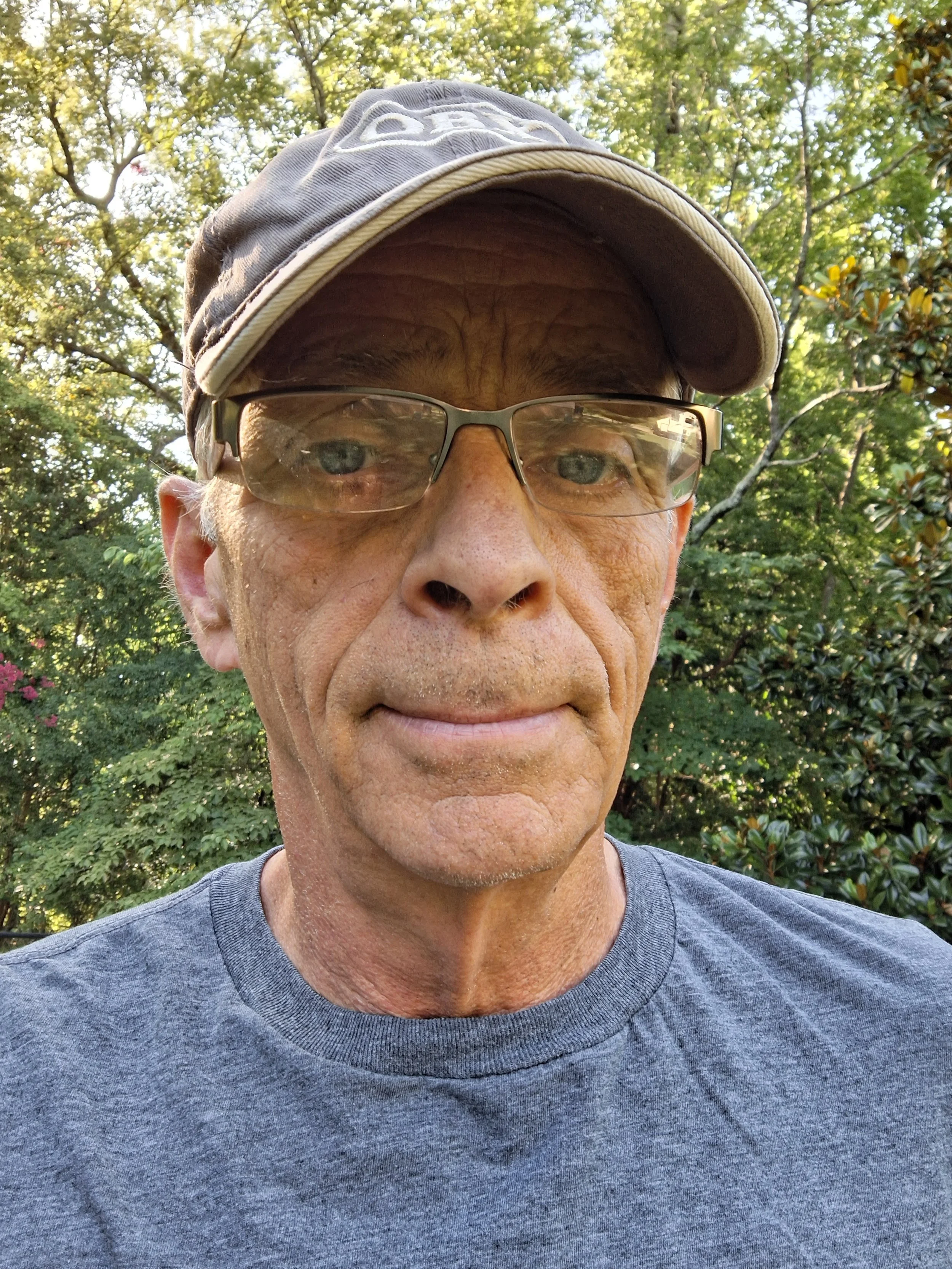 Close-up of a middle-aged man wearing glasses and a baseball cap, standing outdoors with trees in the background.