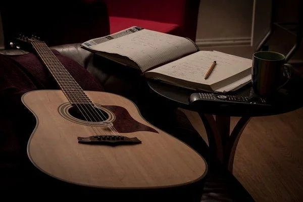 An acoustic guitar placed on a couch next to an open notebook with handwritten notes, a pen, a remote control, and a coffee mug on a small round table in a cozy room.