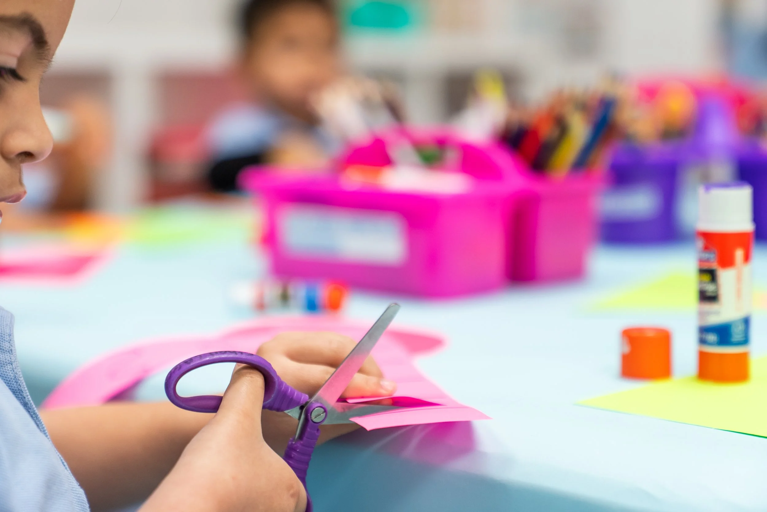 A child with a purple pair of scissors cutting a pink paper at a table surrounded by craft supplies, including glue bottles, markers, and colored paper.