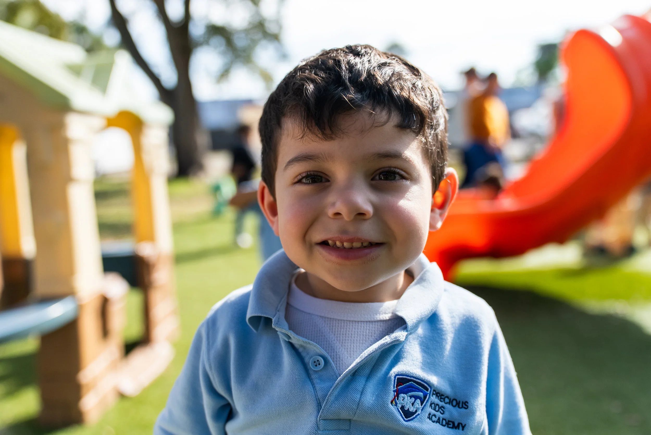 Close-up of a smiling young boy with dark hair, wearing a light blue hoodie with a school logo, outdoors in a park with playground equipment and trees in the background.
