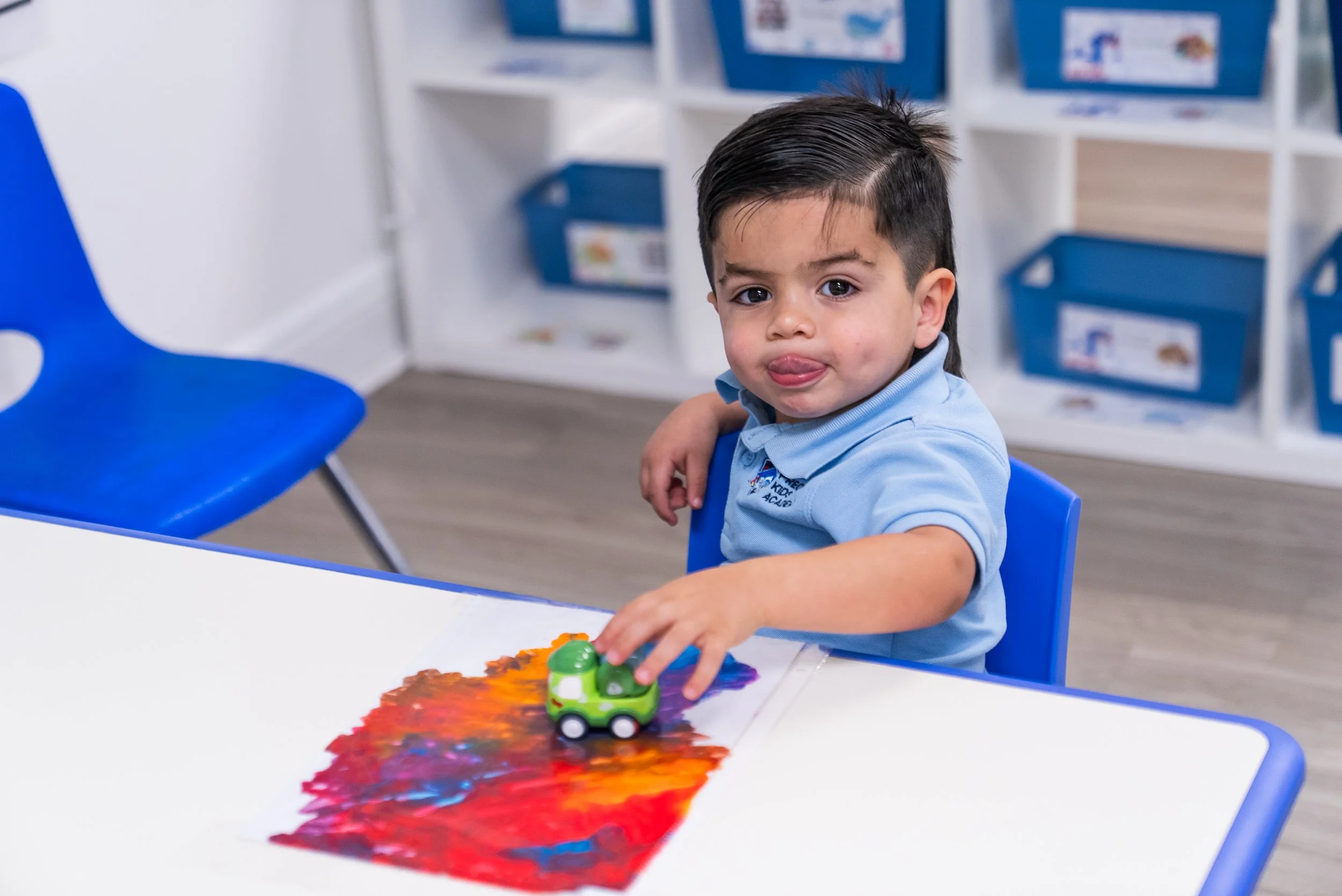 Young boy playing with a toy car on a colorful arts and crafts project at a table in a classroom setting.