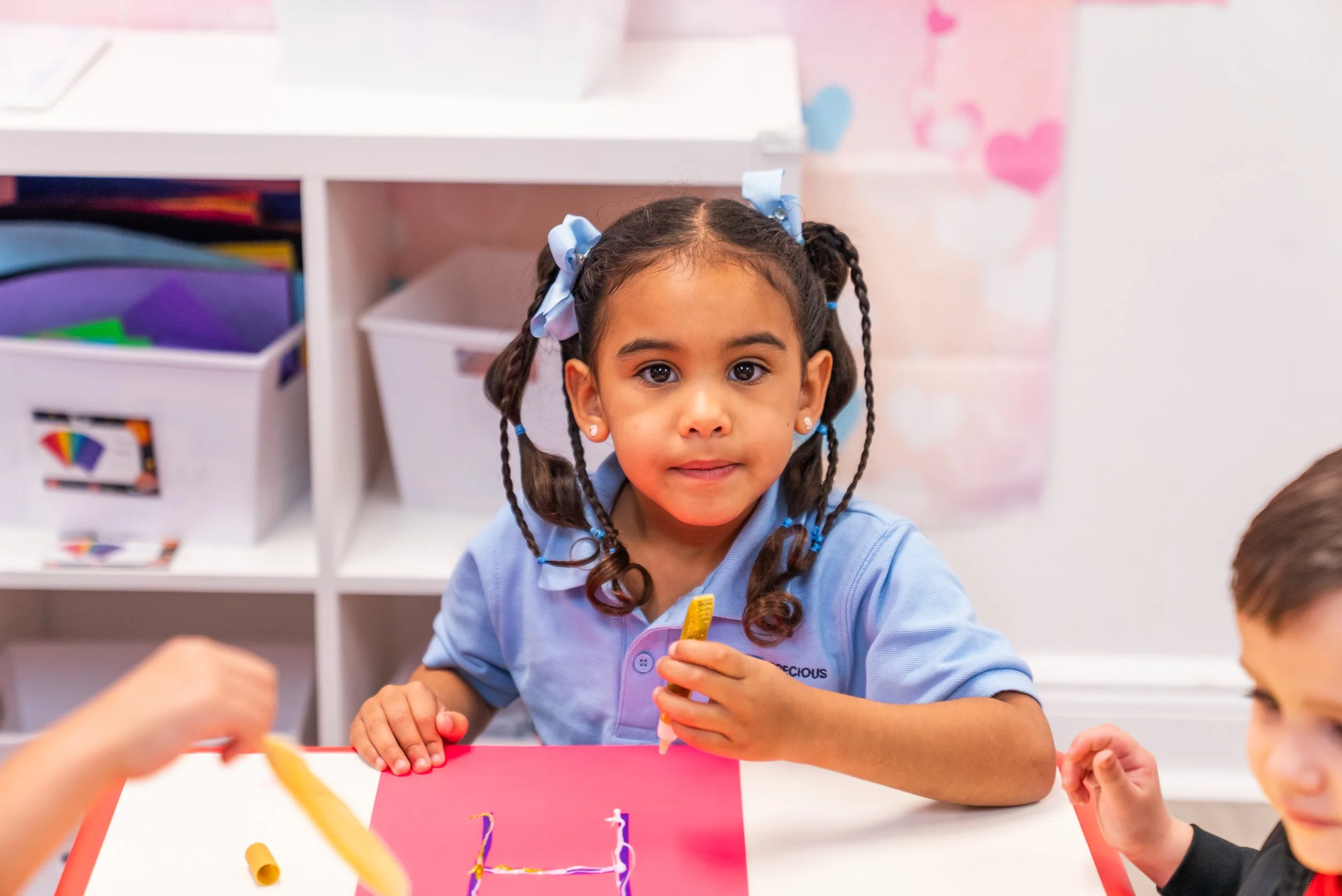 Young girl with braided hair and bows working on a craft project at a table in a classroom.