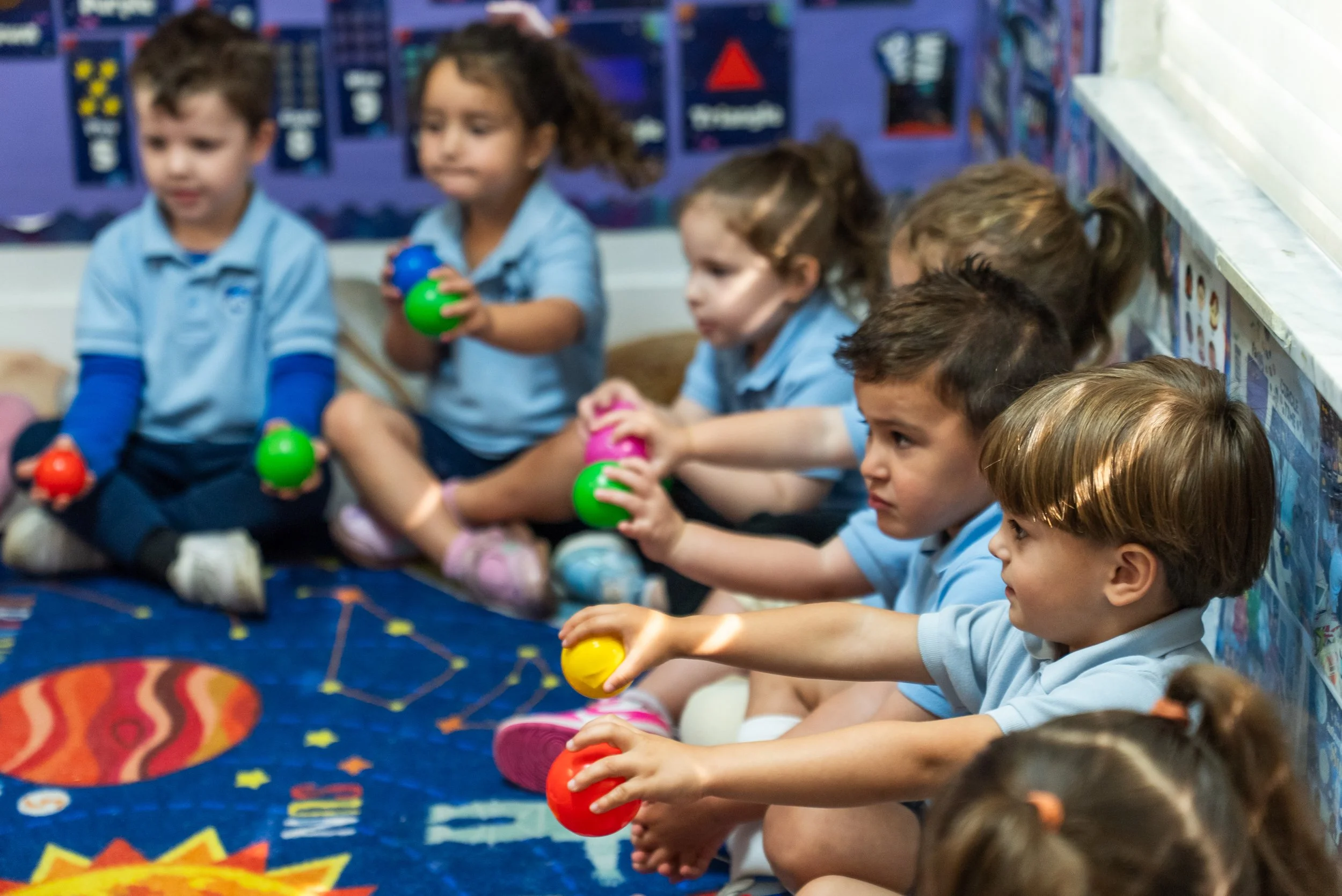 A group of young children sitting on a colorful carpet, holding colorful plastic maracas, in a classroom decorated with educational posters and artwork.