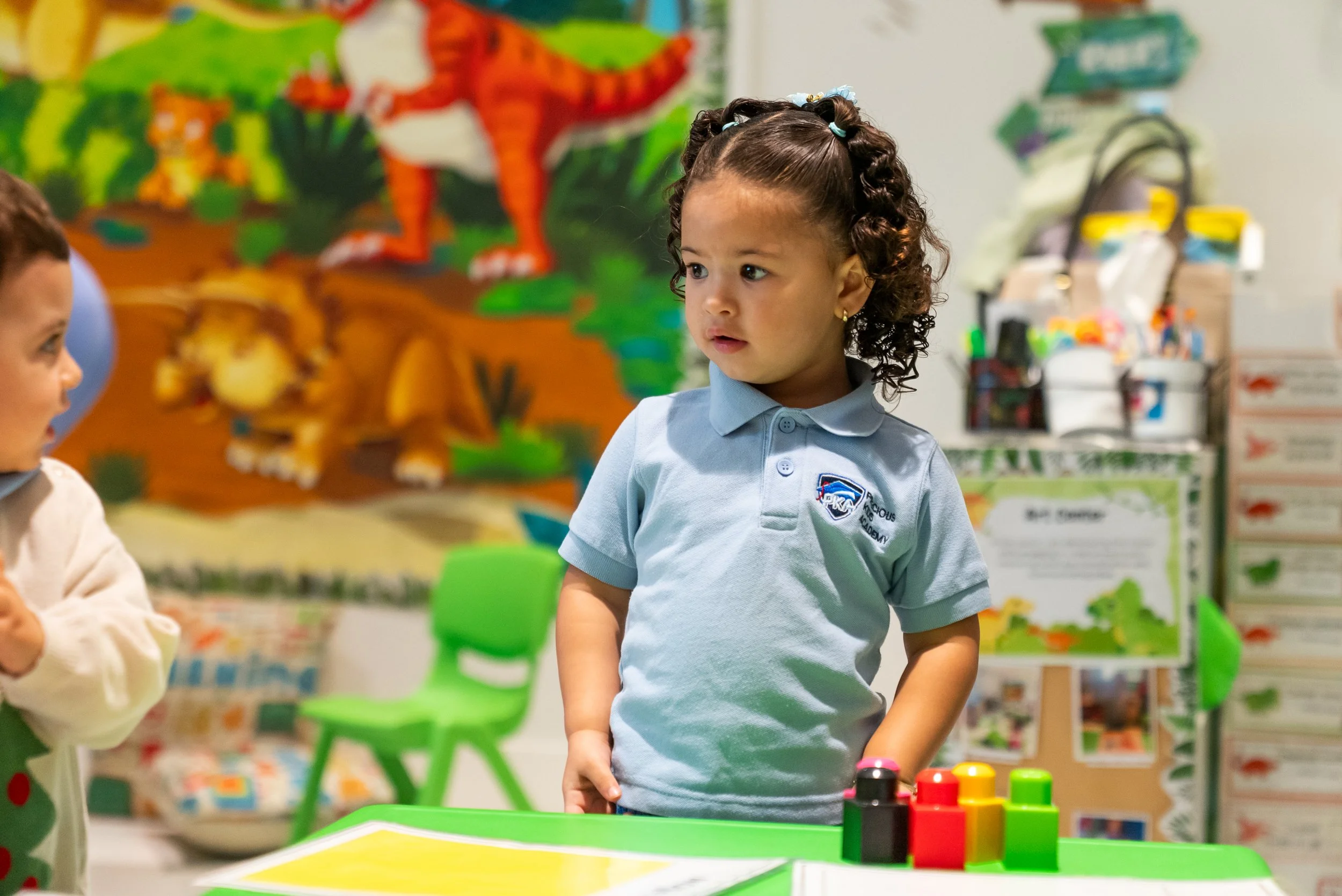 A young girl with curly hair in a ponytail wearing a light blue polo shirt, standing in a classroom with colorful educational decorations and toys. She appears to be engaged in an activity, with a focus on the colorful stacking blocks in front of her.