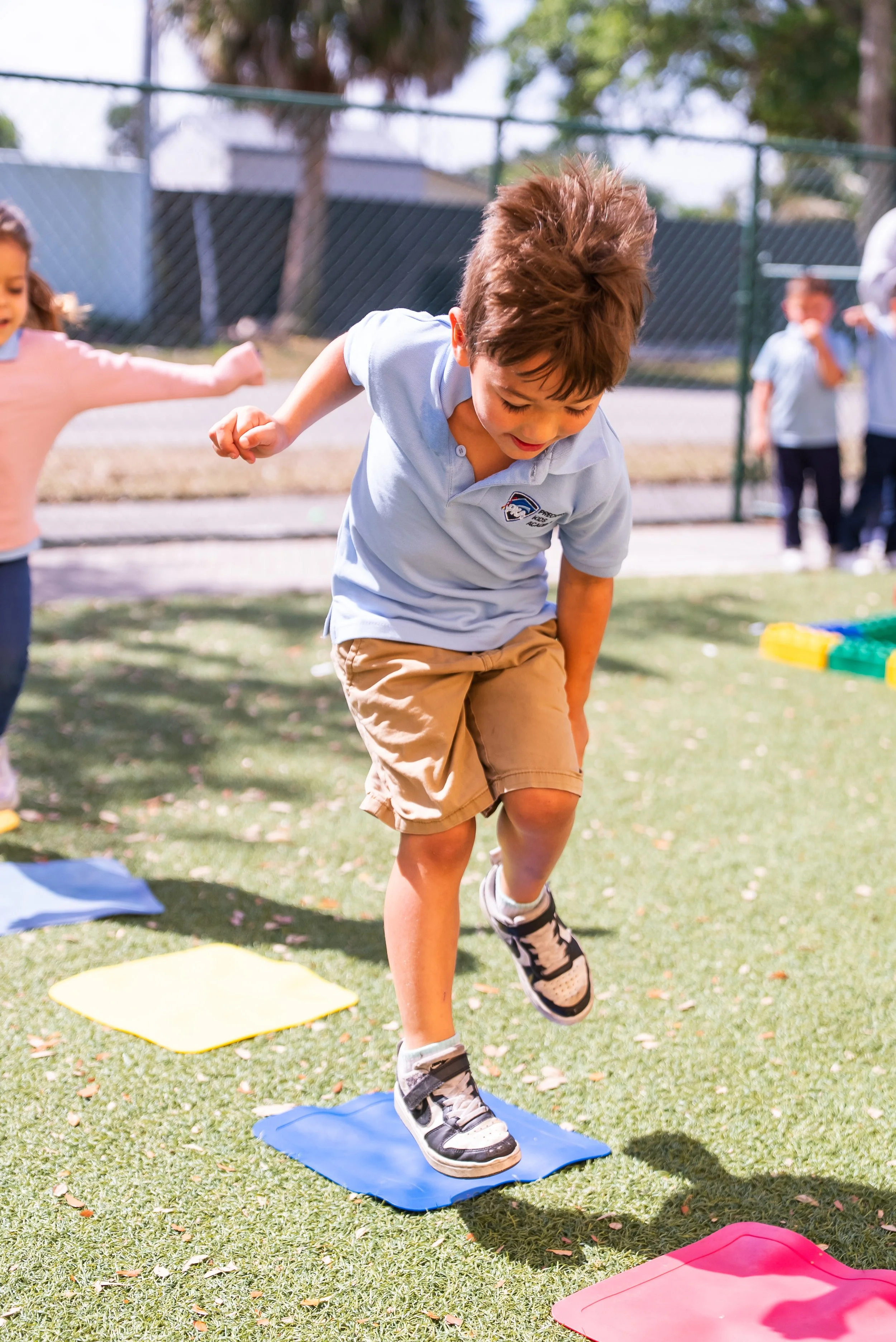 A young boy with brown hair and a light blue polo shirt balancing on a blue agility mat outdoors, with other children and colorful mats in the background.