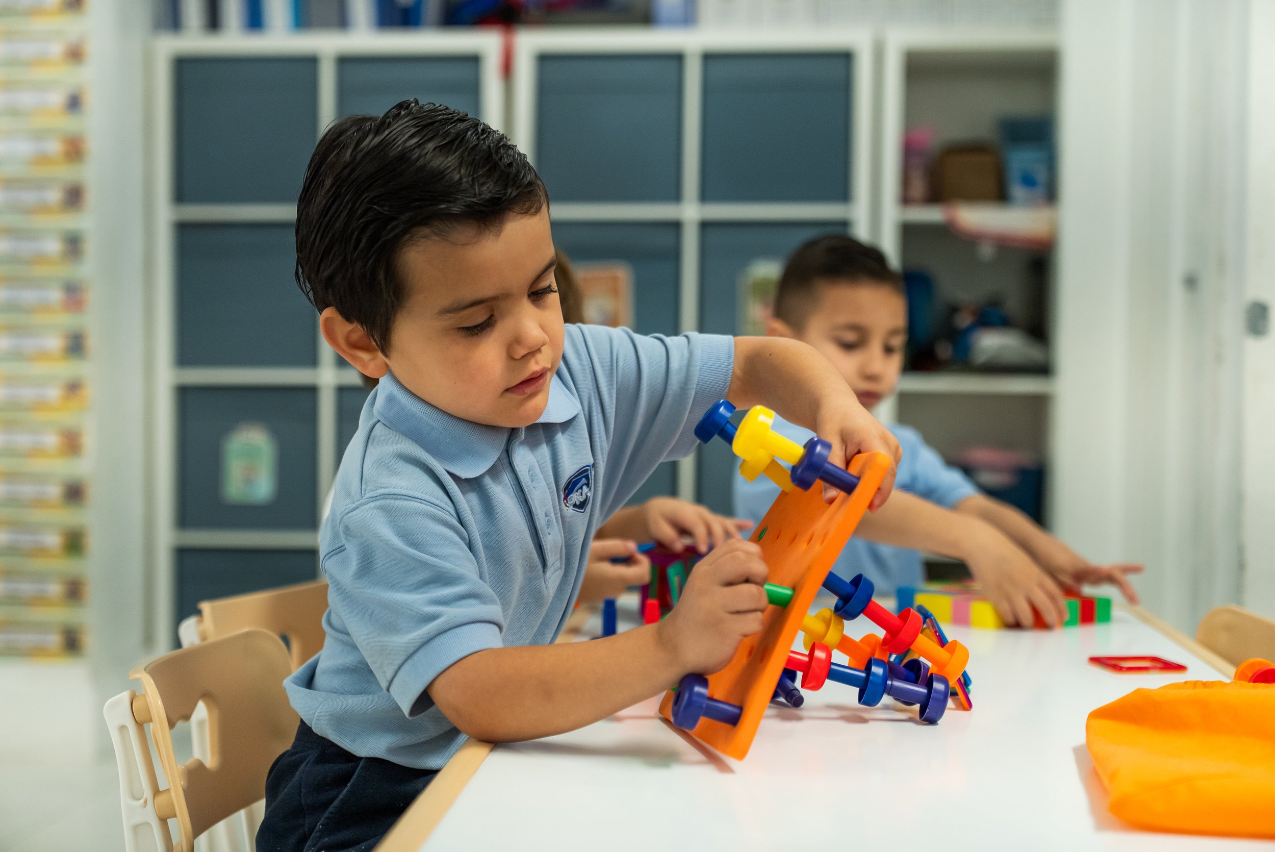 A young boy with dark hair wearing a light blue school uniform working on a colorful educational toy at a table in a classroom. In the background, other children are also engaged in similar activities with shelves and classroom materials visible.