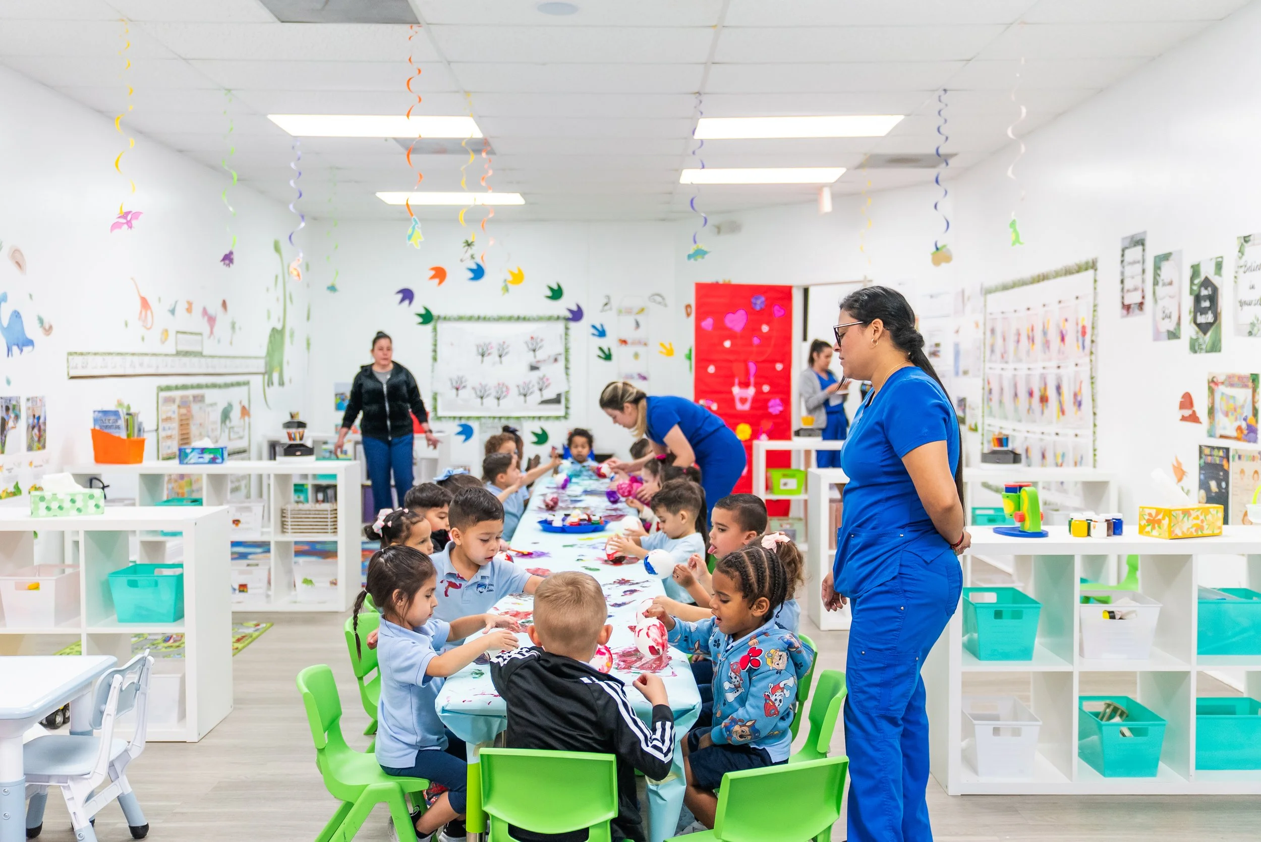Children seated at a long table engaging in arts and crafts activities, with caregivers assisting them, in a brightly decorated classroom with colorful hanging decorations and educational posters.