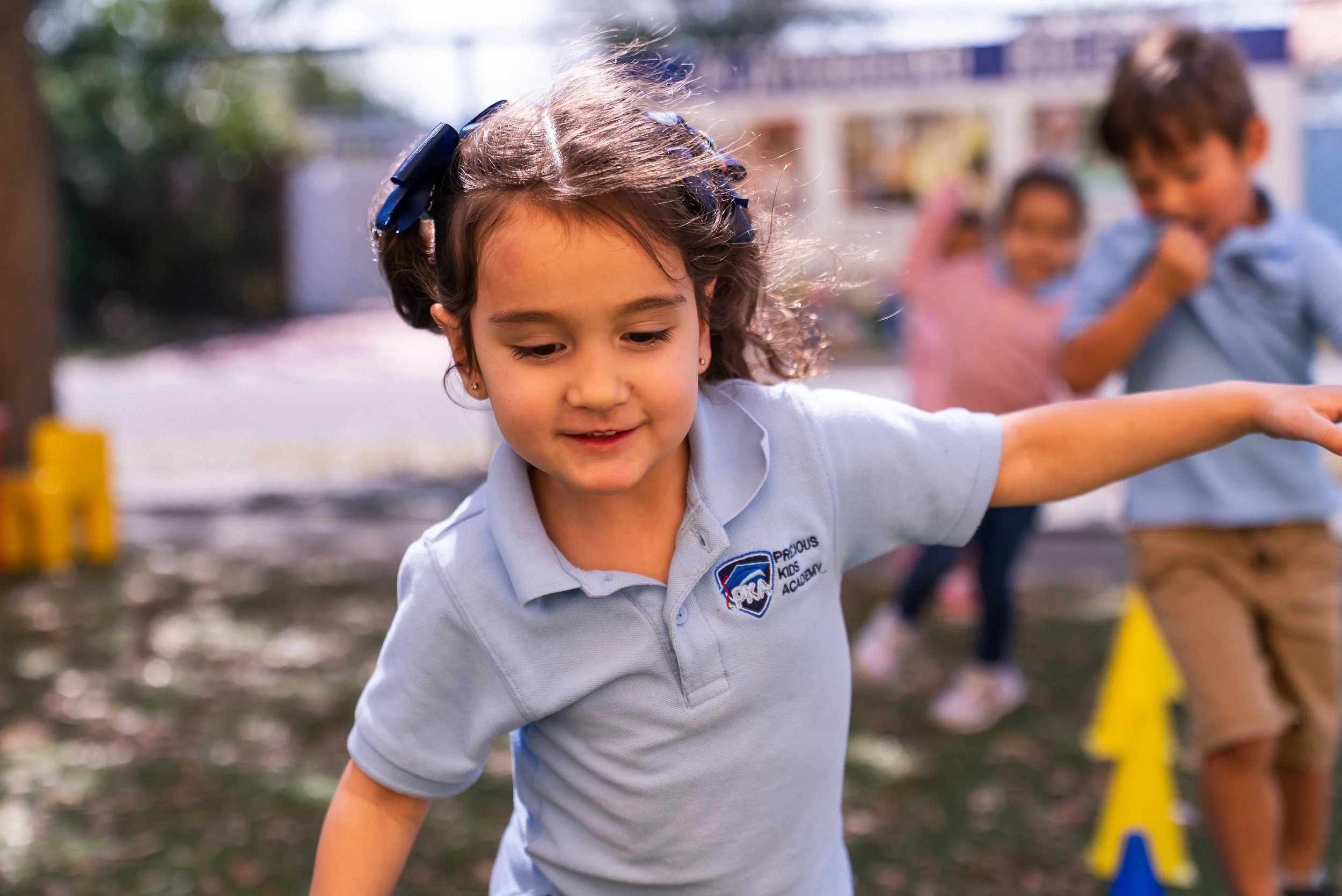Young girl in a school uniform participating in an outdoor activity, with blurred children and playground equipment in the background.