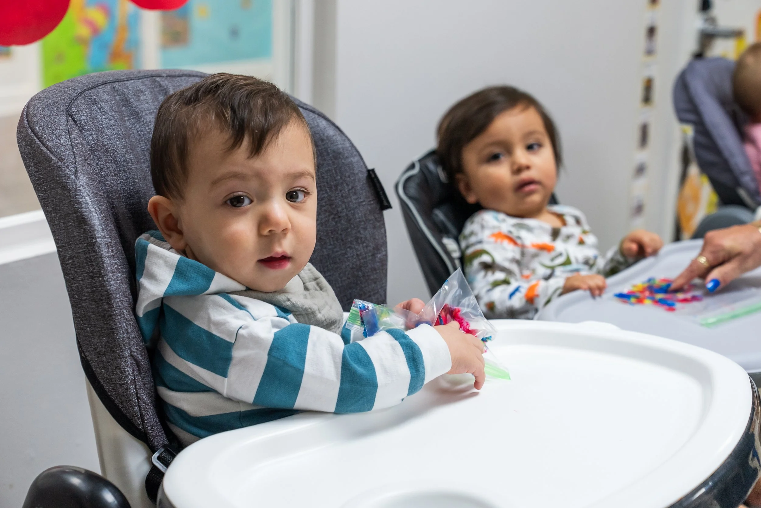 Two young boys sitting in high chairs at a table, engaged in an activity involving colorful craft supplies, in a room with colorful decorations and a white wall.