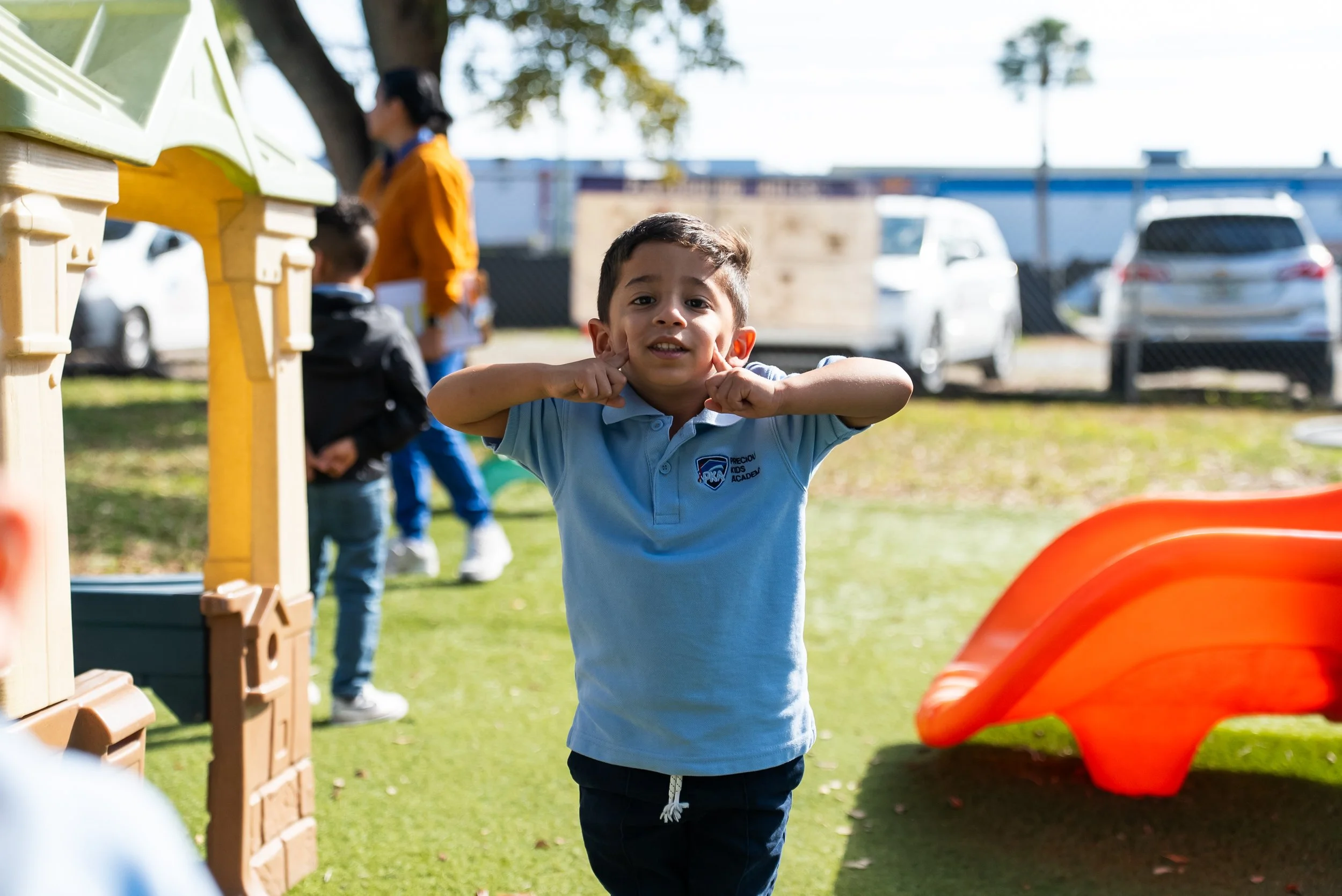 A young boy making a funny face with his hands on his cheeks at an outdoor playground, with other children and a woman in the background.