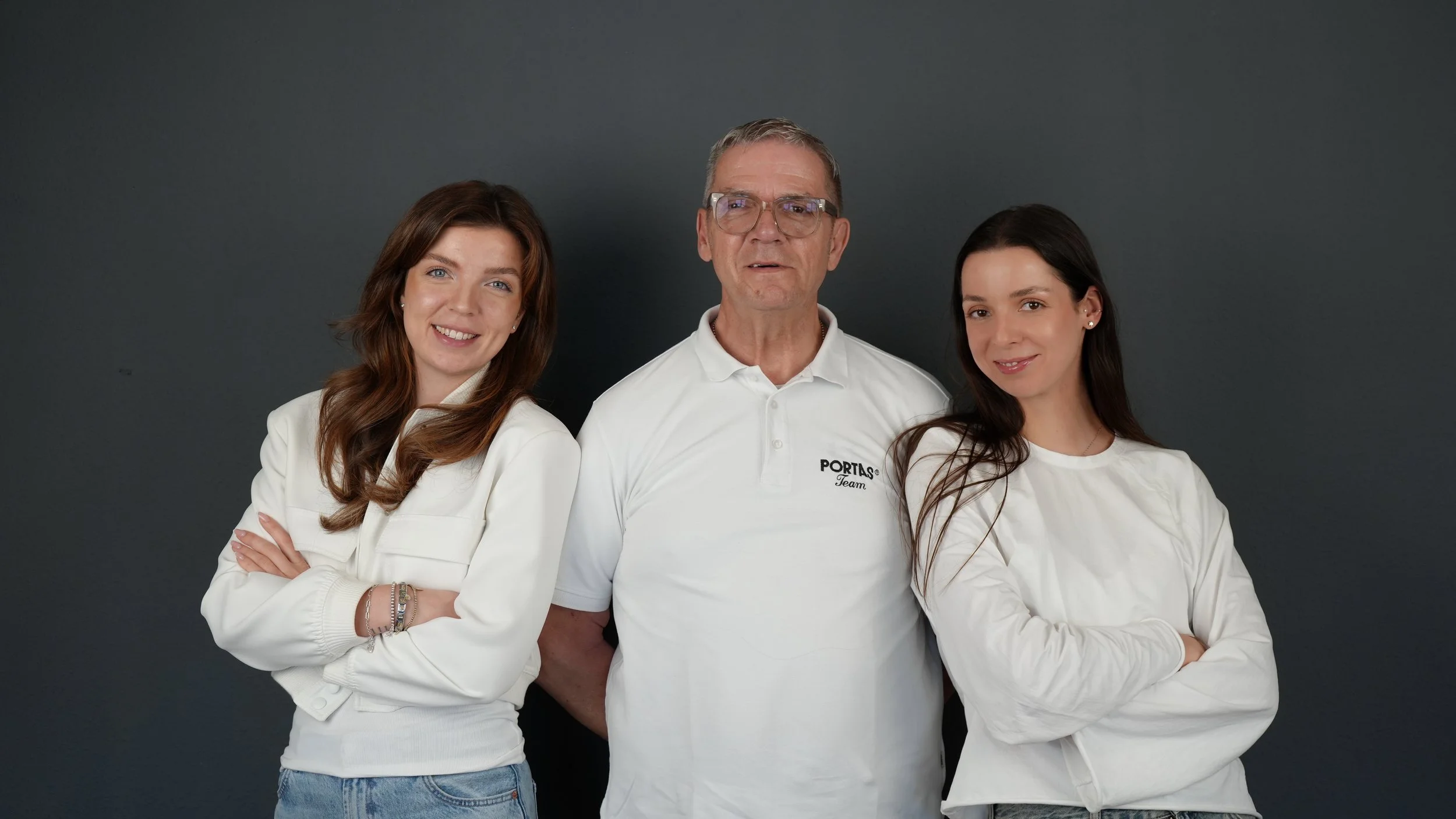 Drei Personen, zwei Frauen und ein Mann, posieren vor einem grauen Hintergrund, alle tragen weiße Oberteile.