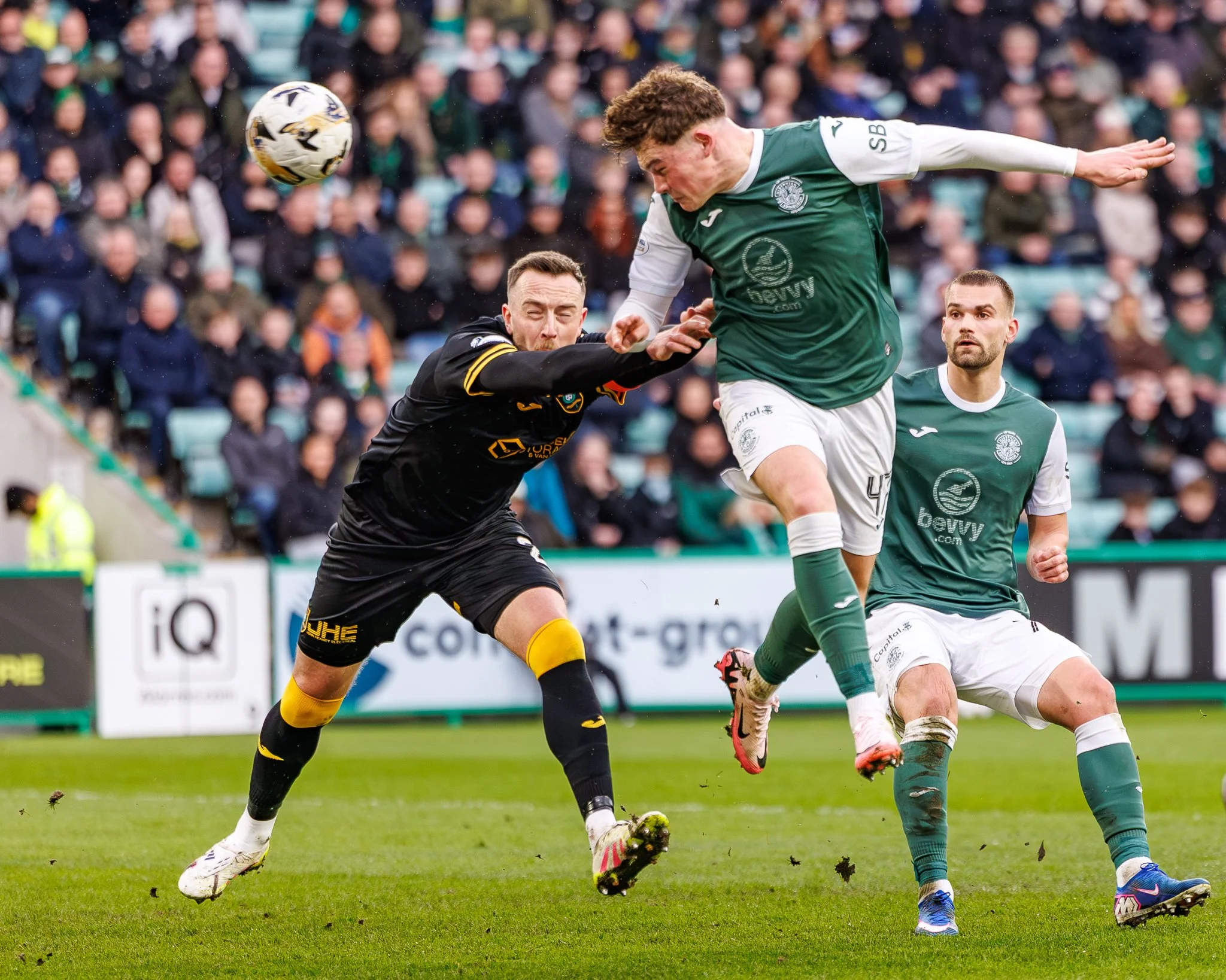 Two soccer players competing for the ball during a match, with one player in a black and yellow uniform and the other in a green and white uniform, with a crowd watching in the background.