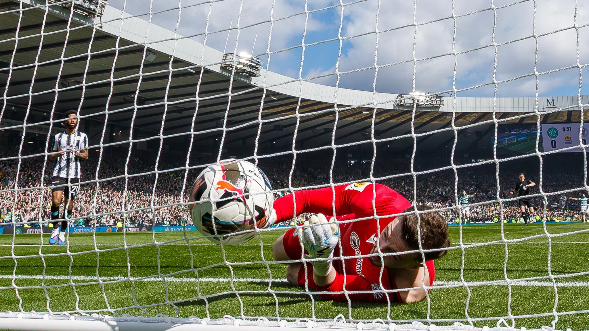 Soccer match scene with a player in black and white striped jersey standing near the goal, a goalkeeper in red diving to save the ball, and a ball heading into the net. Spectators fill the stadium in the background on a partly cloudy day.