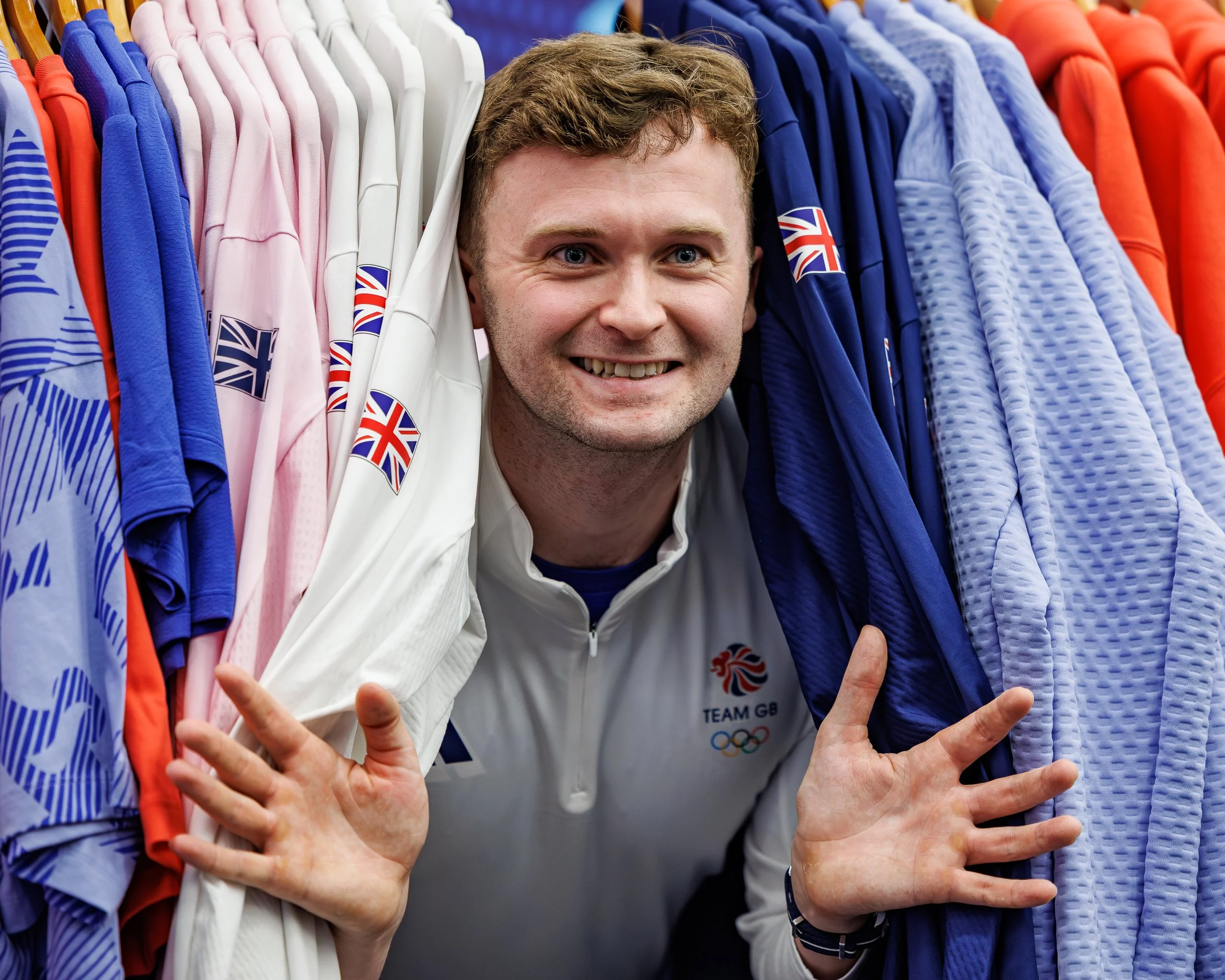 A smiling man wearing a Team GB jacket, surrounded by colorful clothing and waving with multiple hands.