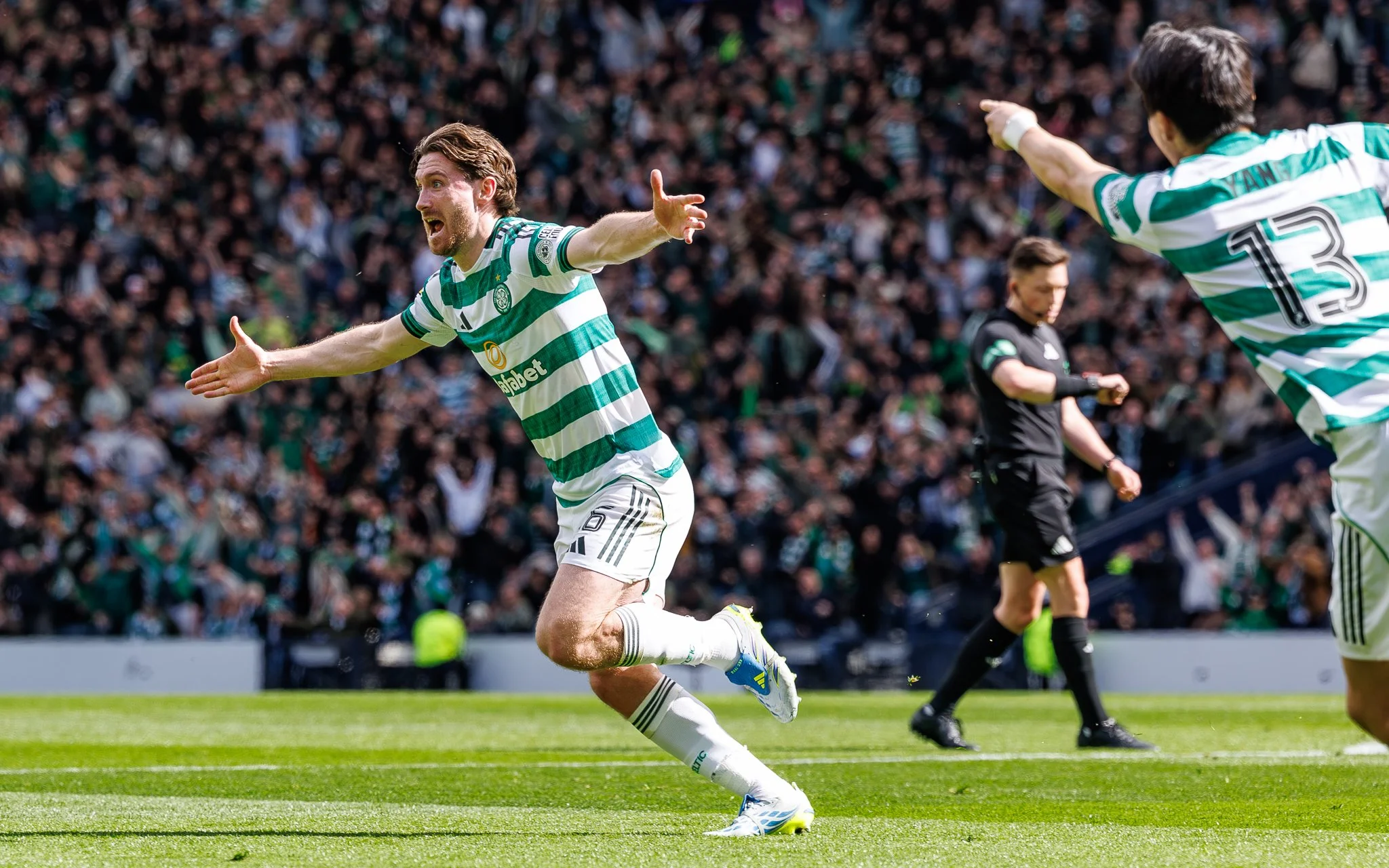 A soccer player is running across a green field celebrating, wearing a green and white striped jersey with the number 13, with a crowd of spectators in the background.