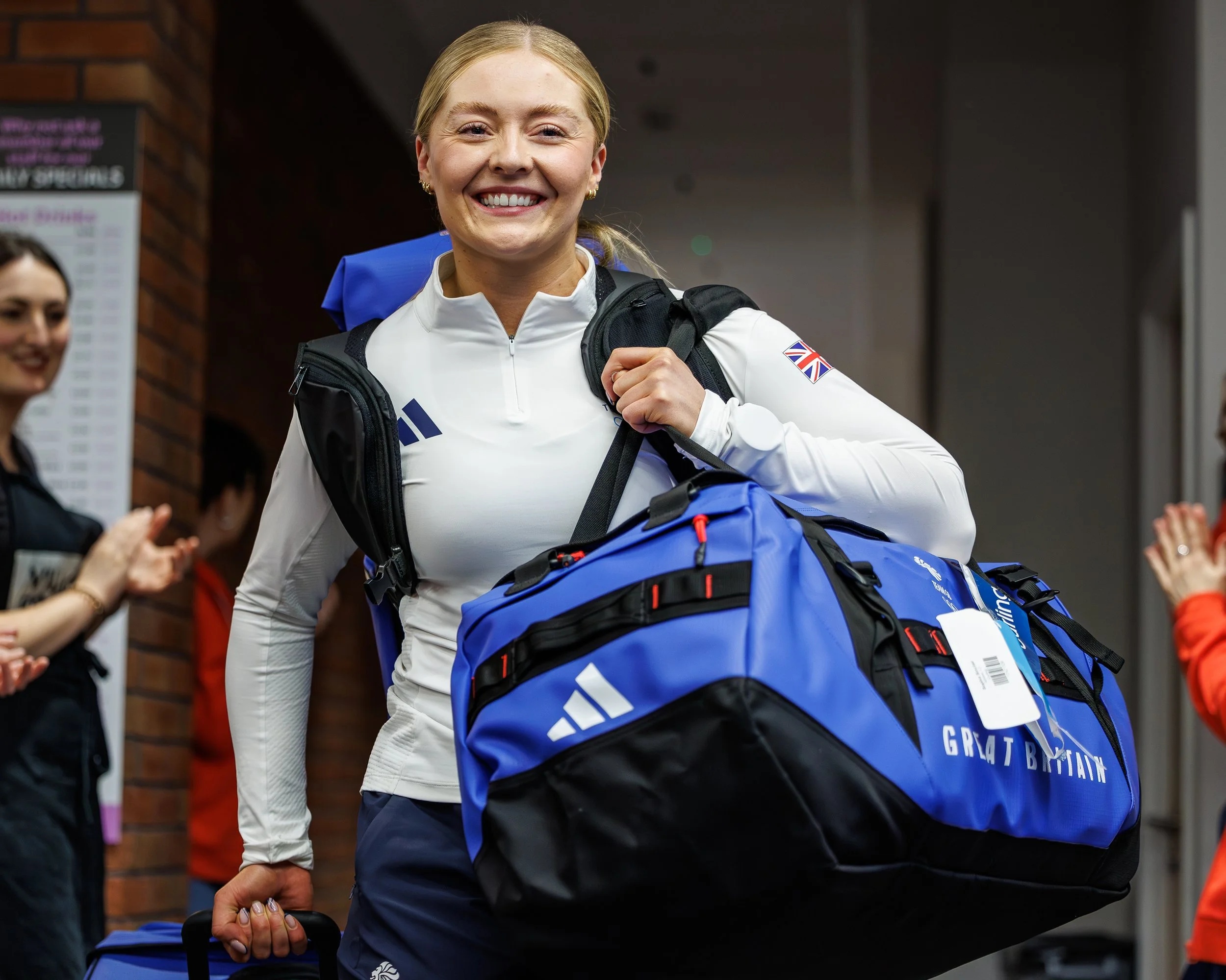 Smiling woman with blonde hair, wearing a white sports jacket with a Union Jack patch, carrying a blue travel bag, at an indoor event with onlookers.