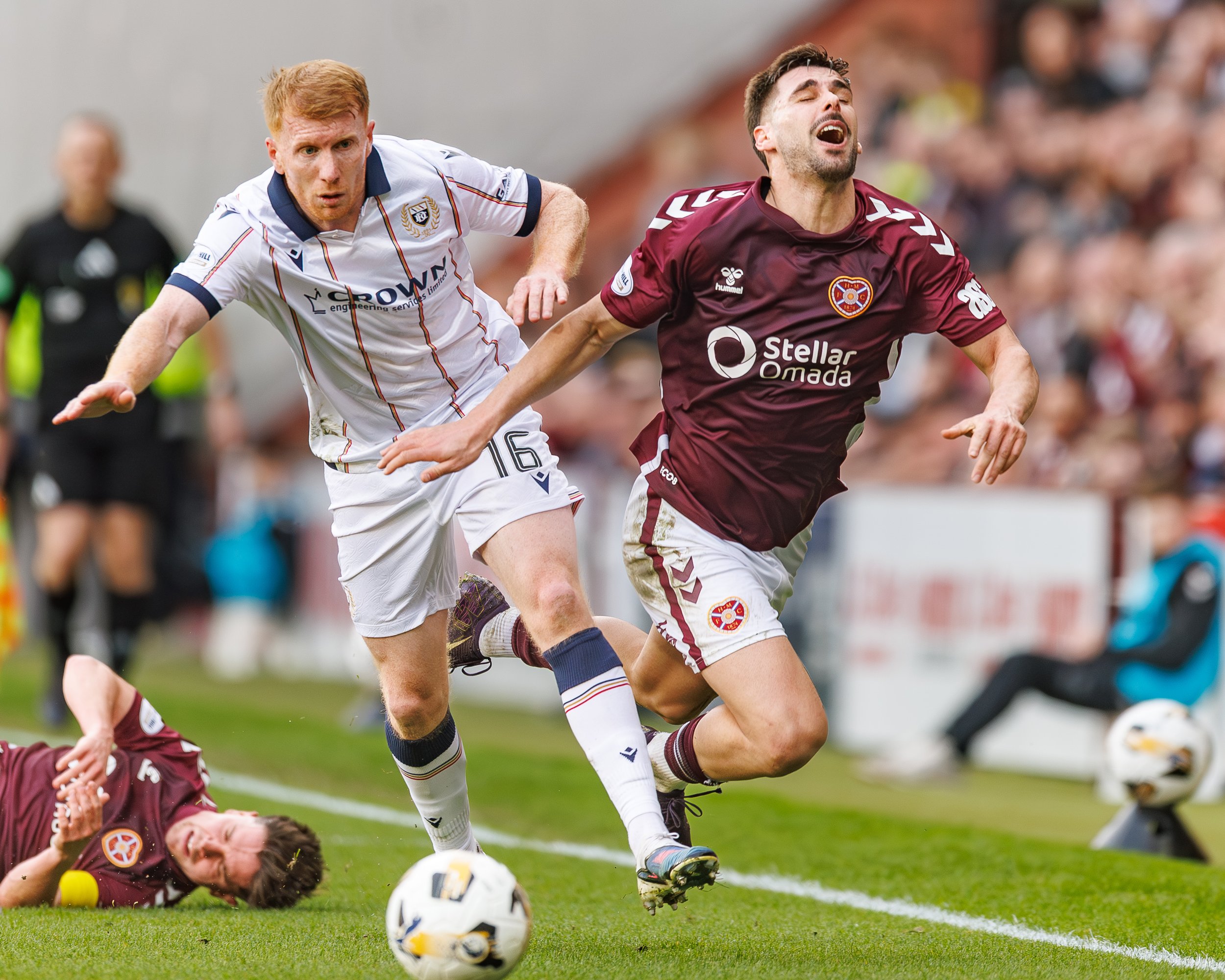 Two soccer players racing for the ball on the field, one in a white jersey and the other in a maroon jersey, with a blurred crowd in the background.