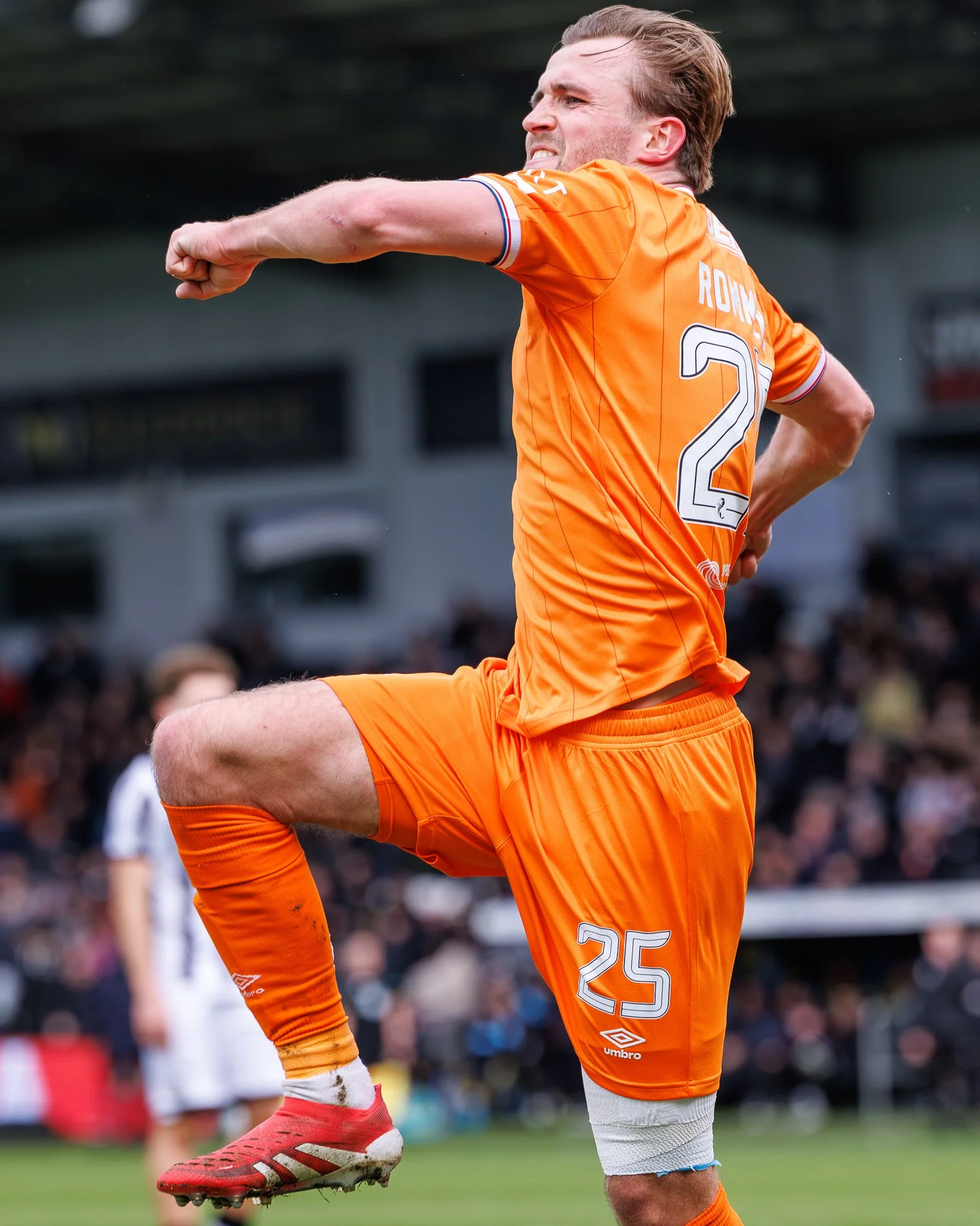 A soccer player in an orange uniform is in motion, possibly celebrating or stretching on the field, with a blurred crowd in the background.