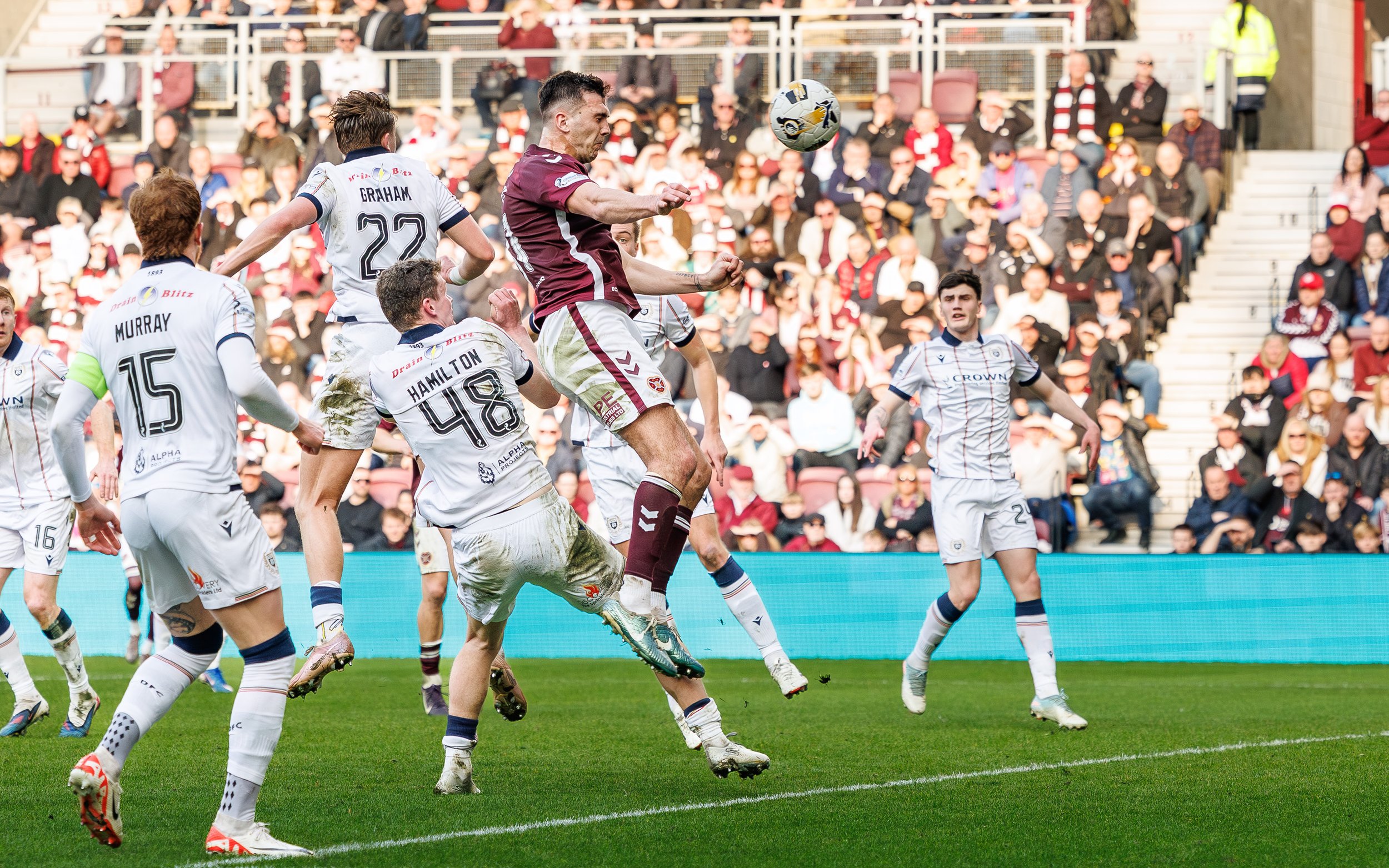 A soccer match in progress with players in white and maroon uniforms. One player is jumping to head the ball while others are on the field. Spectators are in the stands watching the game.