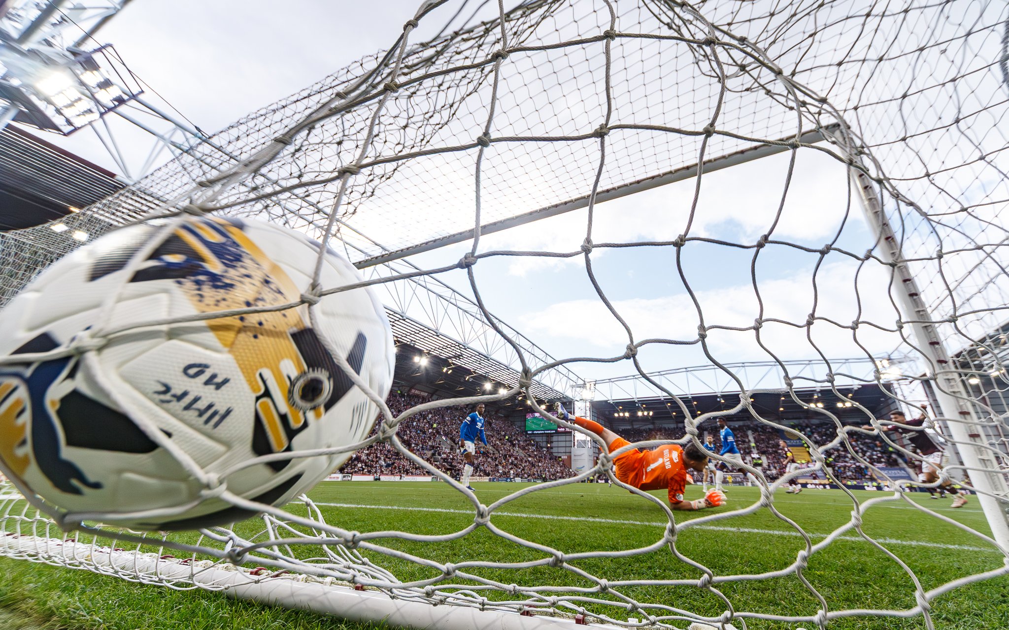 A soccer ball is seen hitting the net of the goal during a match. In the background, players are on the field, and crowd-filled stands are visible at a stadium on a cloudy day.
