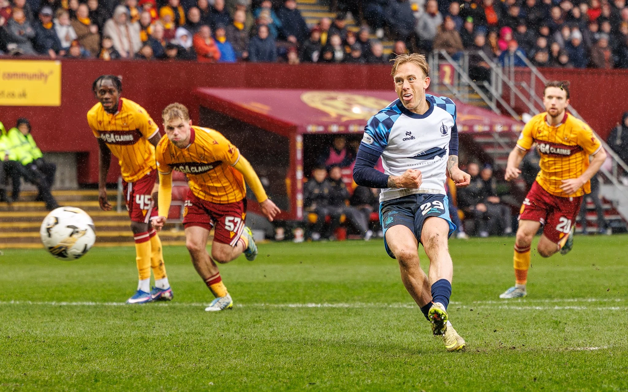 A soccer game in progress with players running after the ball on a wet field, with a crowd in the stands.