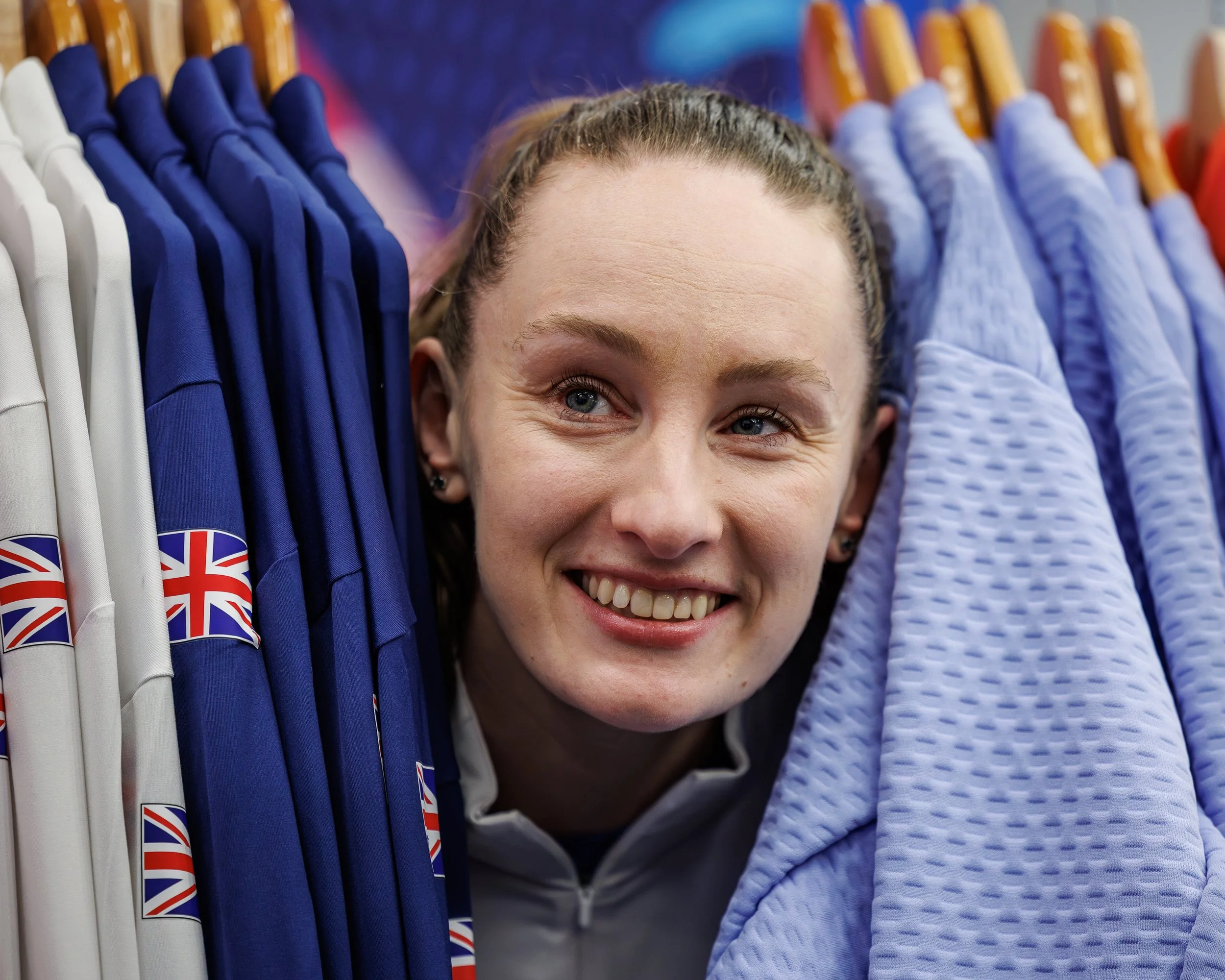 A woman smiling with her face between clothing items such as t-shirts and jackets on hangers, mainly in blue and white, with some British flags visible on the clothing.