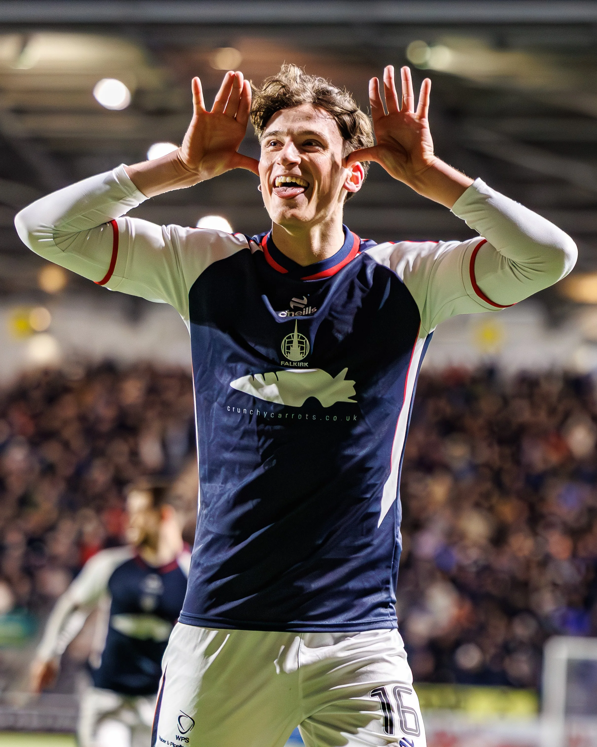A jubilant soccer player in a white and navy jersey with red accents, standing on a soccer field, celebrating with hands near his ears, sticking out his tongue, and smiling.
