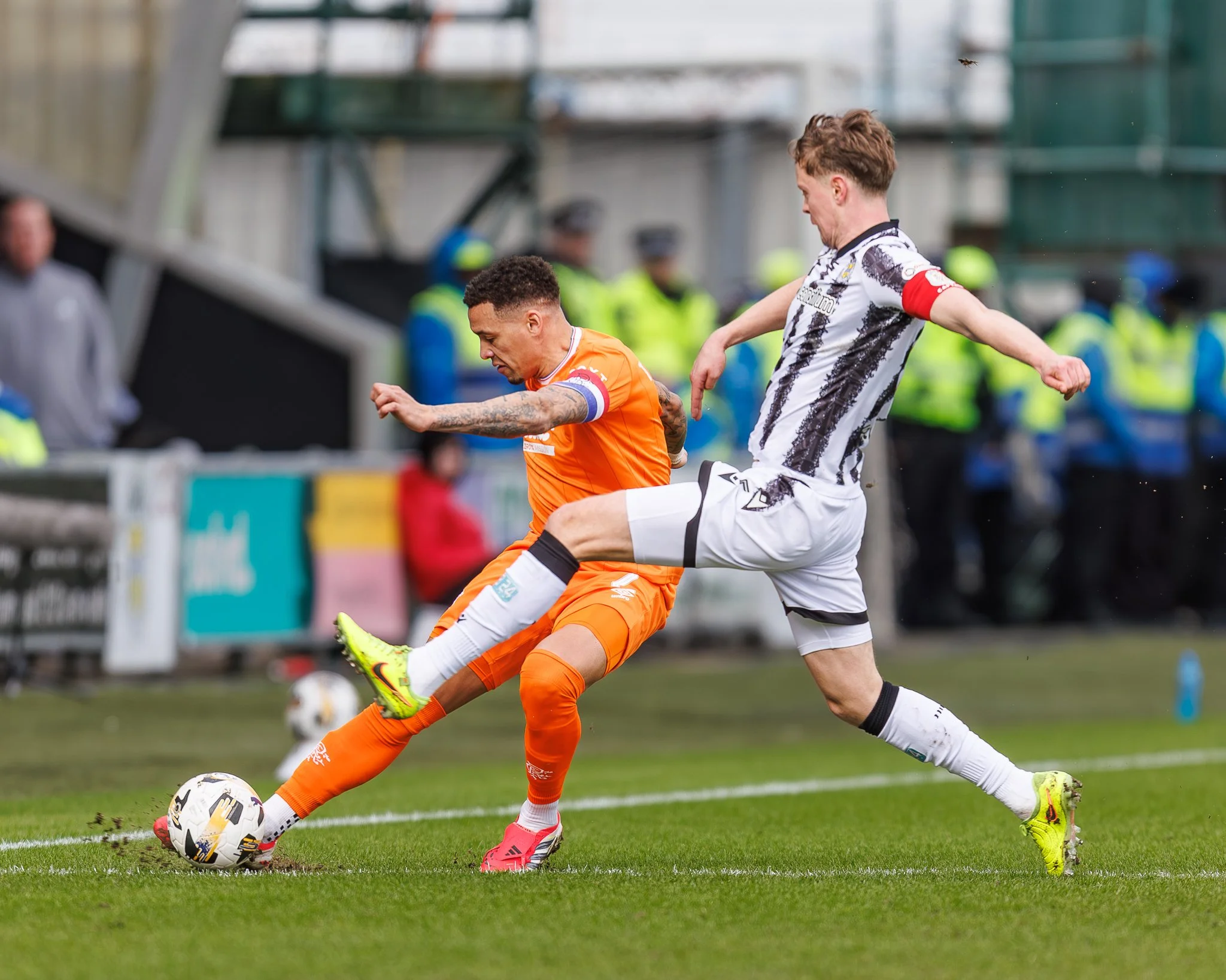 Two soccer players compete for the ball during a match, with spectators and security in the background.