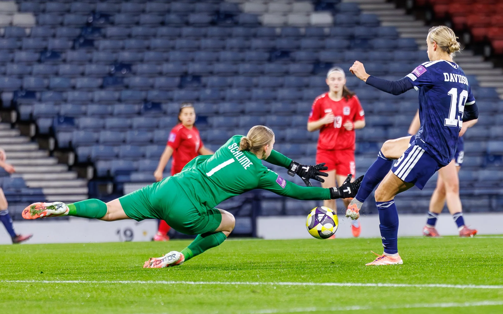 A soccer player in a blue uniform kicks a ball while a goalkeeper in a green uniform dives to intercept the shot on goal. Two other players in red uniforms observe in the background, along with empty stadium seats.