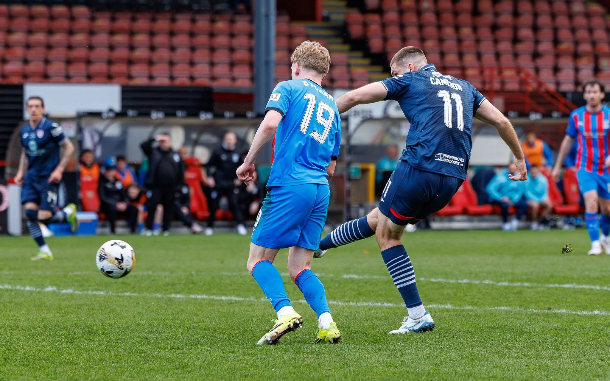 Two soccer players in blue jerseys compete for the ball during a match on a green field, with other players and spectators in the background.