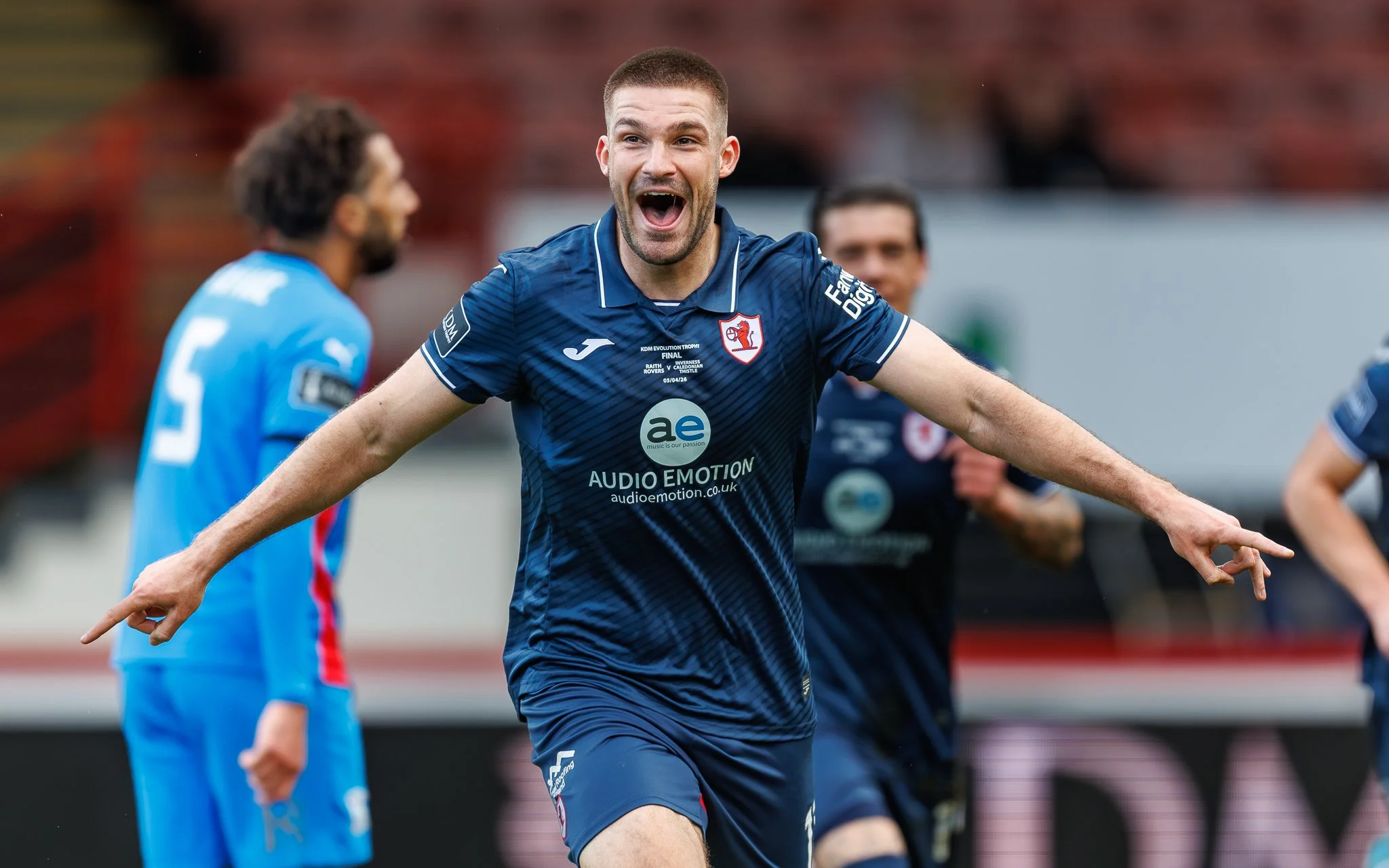 A man in a blue sports jersey celebrates during a soccer match, with other players visible in the background.