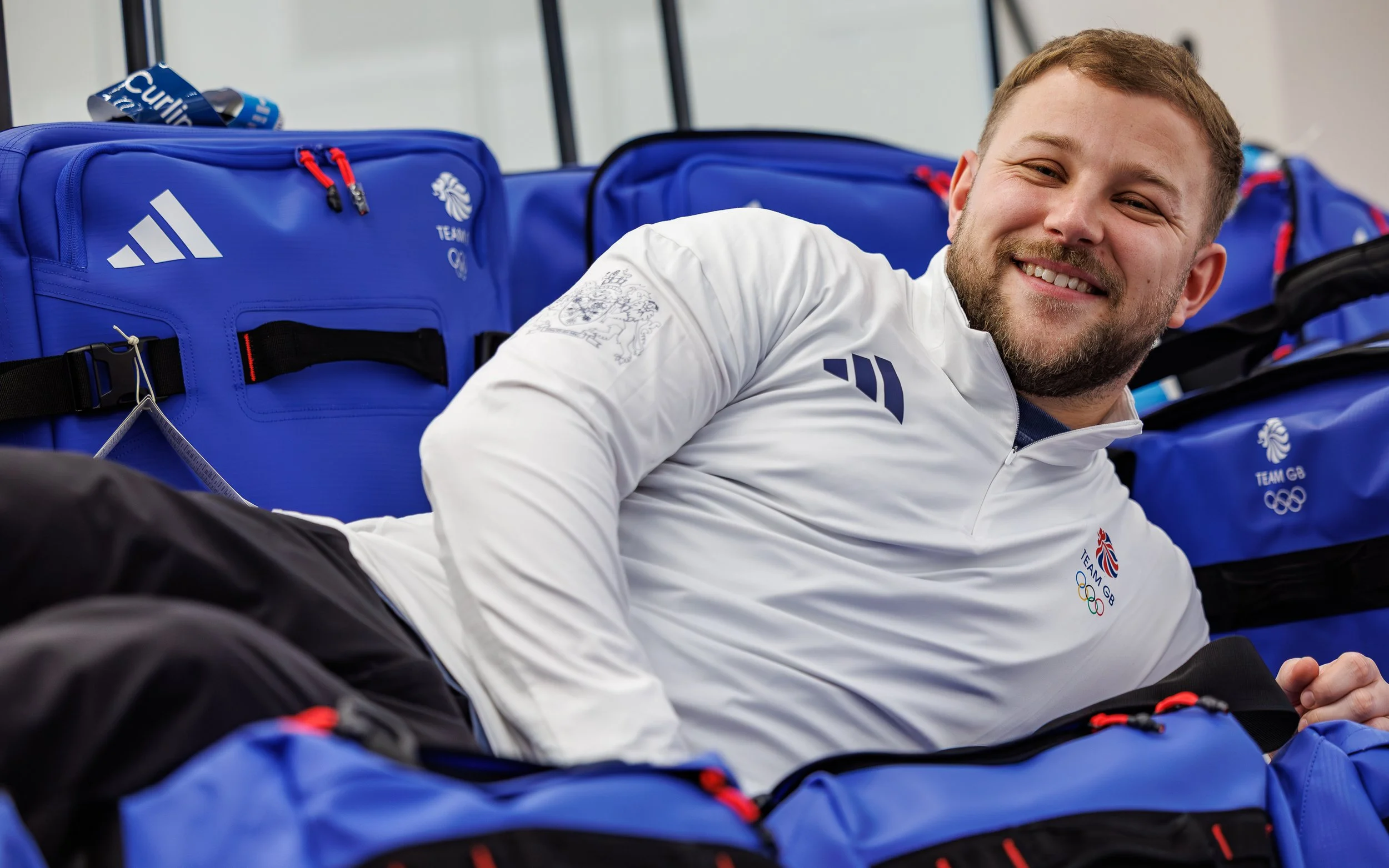 A smiling man in a white Team GB jacket with Olympic logos, lying on blue rolling luggage with red zippers at what appears to be an airport or transportation hub.