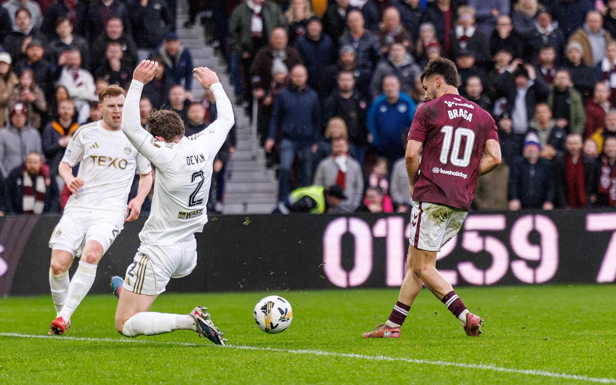 Soccer players in white and maroon uniforms competing for the ball during a match, with a crowd of spectators in the background.